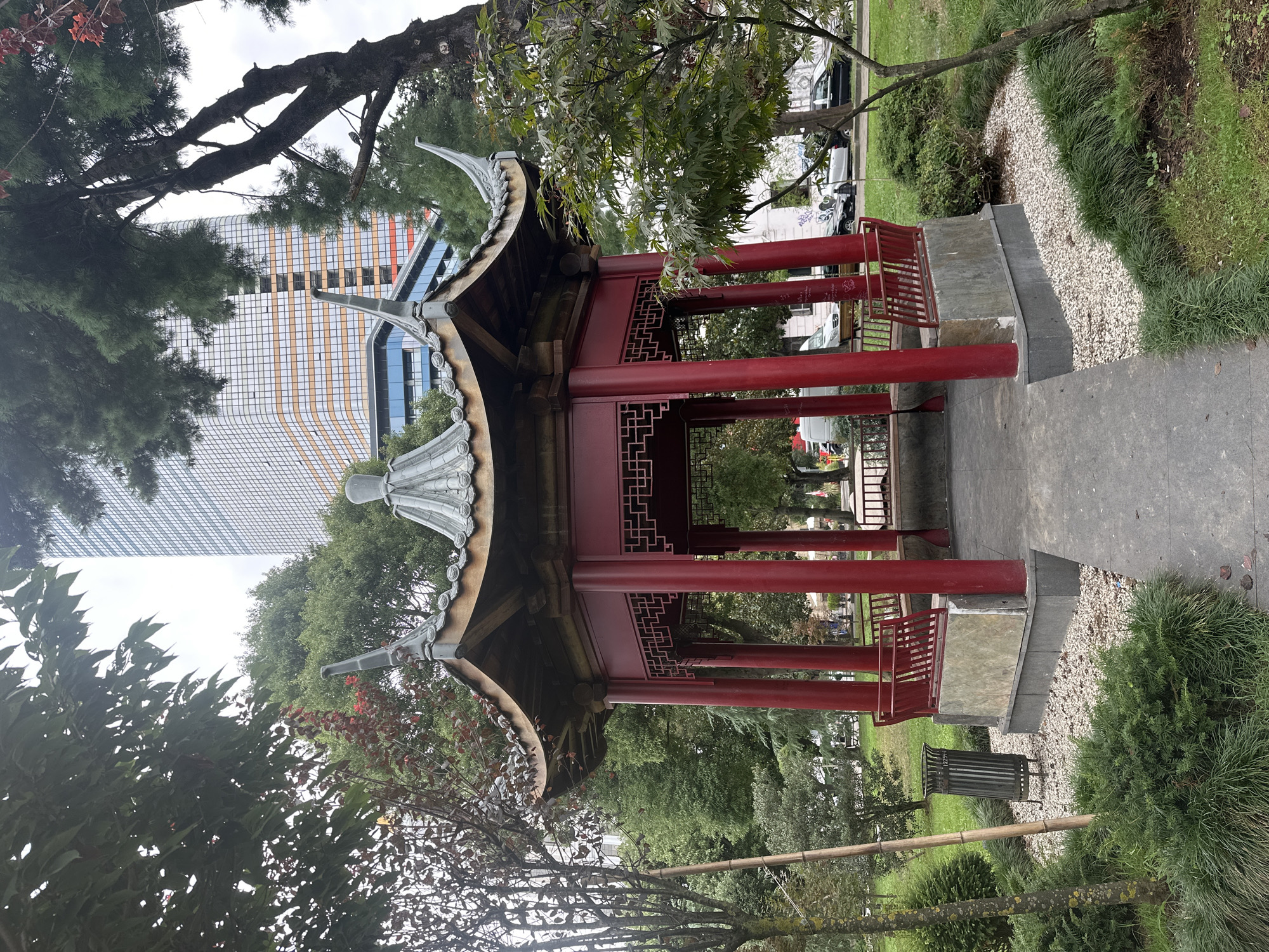 The image shows a traditional red Chinese gazebo in a park in Batumi, Georgia, with a modern skyscraper in the background. The gazebo has a tiered roof with decorative eaves and is supported by red pillars. Inside, there are benches with red railings. The park area around the gazebo is landscaped with green grass, bushes, and small gravel paths. A metal trash can is visible to the left of the gazebo. In the background, a tall, contemporary building with a glass facade and orange accents stands prominently. Cars are parked on a street adjacent to the park. The sky is overcast.