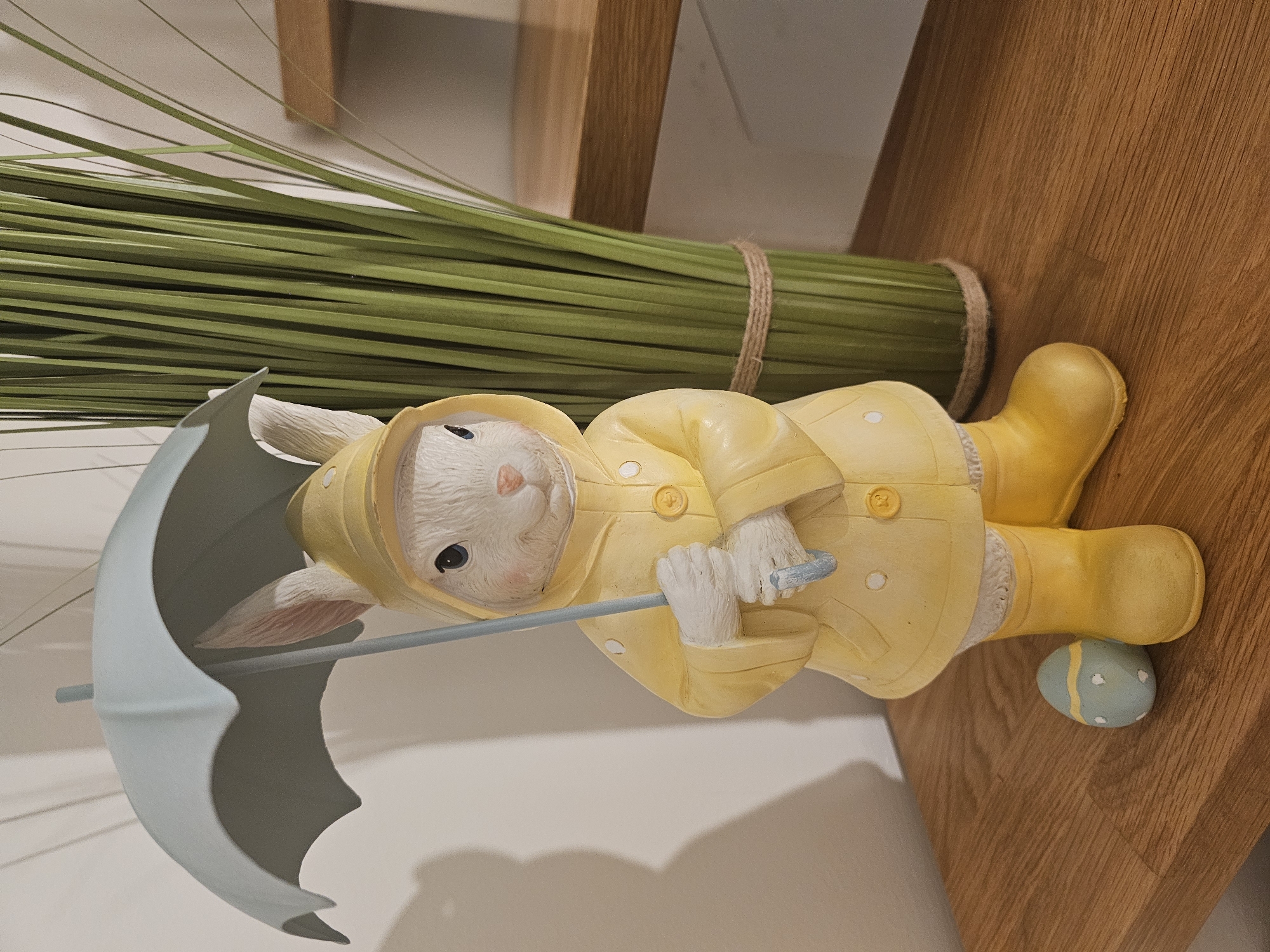 A statue of a rabbit wearing a yellow raincoat and yellow boots stands on a wooden surface, holding a light blue umbrella. The rabbit is looking forward with its paws holding the umbrella. Next to the rabbit's feet is a light blue and yellow striped Easter egg. Behind the rabbit, a bundle of green, grass-like decorations is tied with twine. The background is a plain wall with a shadow cast on it, and a wooden shelf is visible to the right.