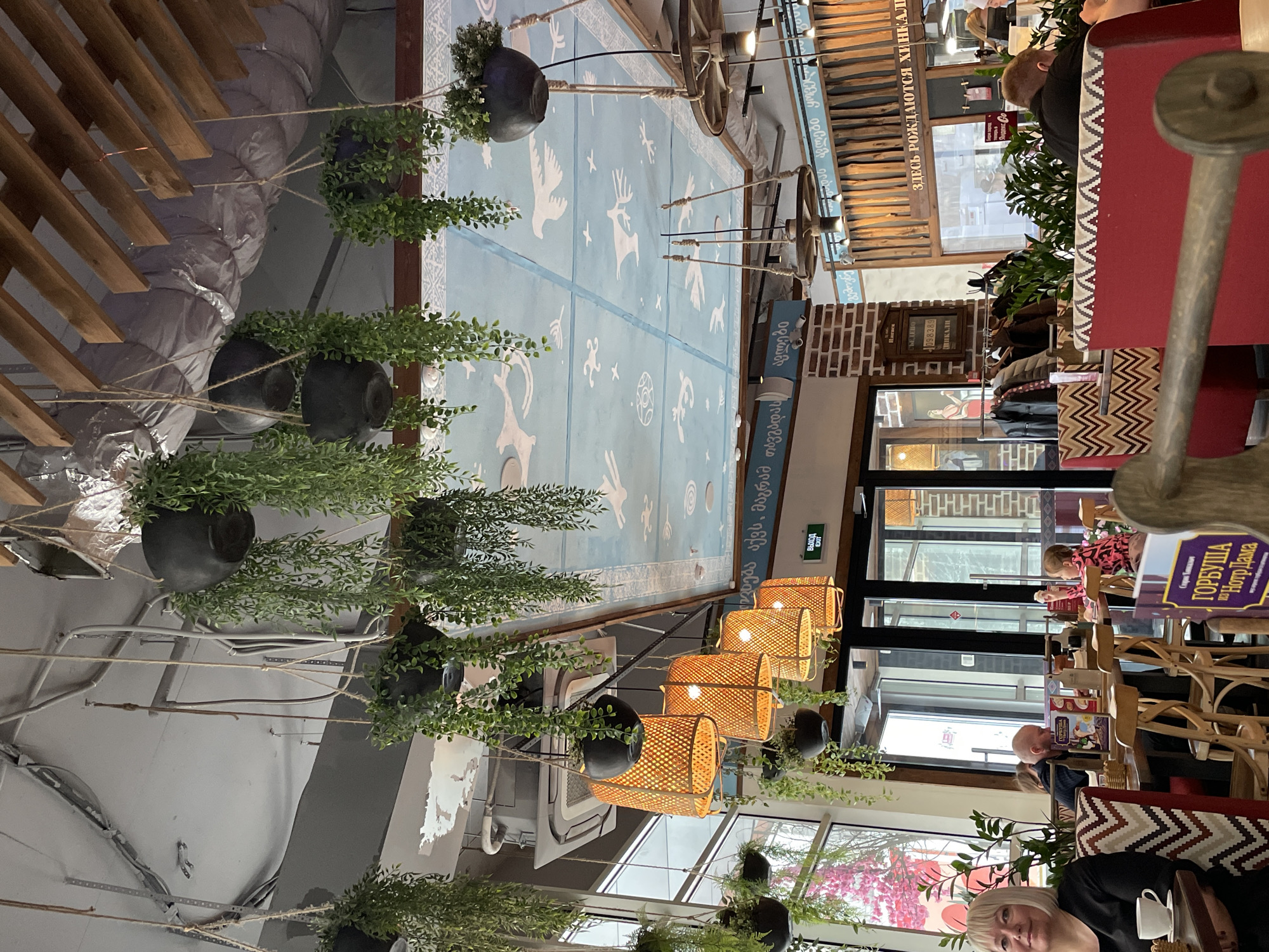 The image shows the interior of a restaurant with decorative elements including hanging plants, woven lanterns, and a ceiling painted with light blue folk motifs. Customers are seated at tables, including one woman holding a white coffee cup. One menu is visible with Cyrillic text. Text in Georgian is also present. The scene is indoors, illuminated by natural light from windows and the hanging lanterns. Some text is in Russian. A sign indicates an exit. The scene is in a restaurant, possibly in Izhevsk, Russia.