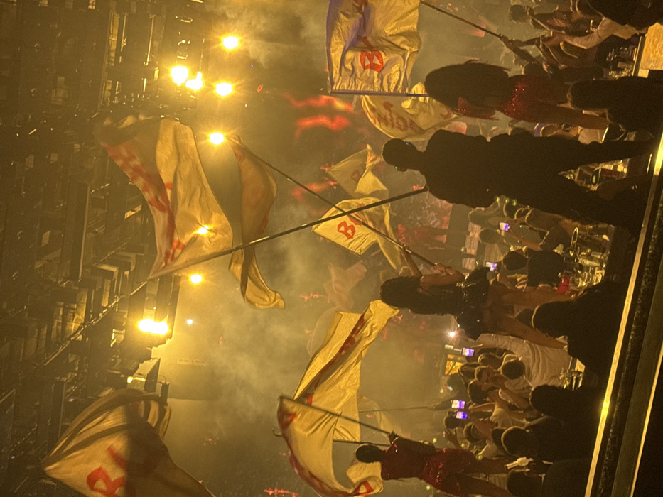 The image captures an active indoor entertainment venue, likely a club or concert hall, within the city of Plovdiv, Bulgaria. The environment is characterized by dim ambient lighting, dense atmospheric haze, and multiple bright yellow and orange spotlights emanating from above. In the foreground, three primary figures (two females, one male) stand on an elevated surface, observed from behind. The female on the left wears a shimmering red outfit, while the central female wears a dark, short-skirted ensemble. The male figure is dressed in dark clothing. All three are actively waving large, rectangular yellow flags featuring a red design, possibly a letter 'B' or 'R'. Below these figures, a large crowd of individuals is visible, many holding up illuminated mobile phones. The immediate foreground shows tables with multiple bottles, glasses, and ice buckets.