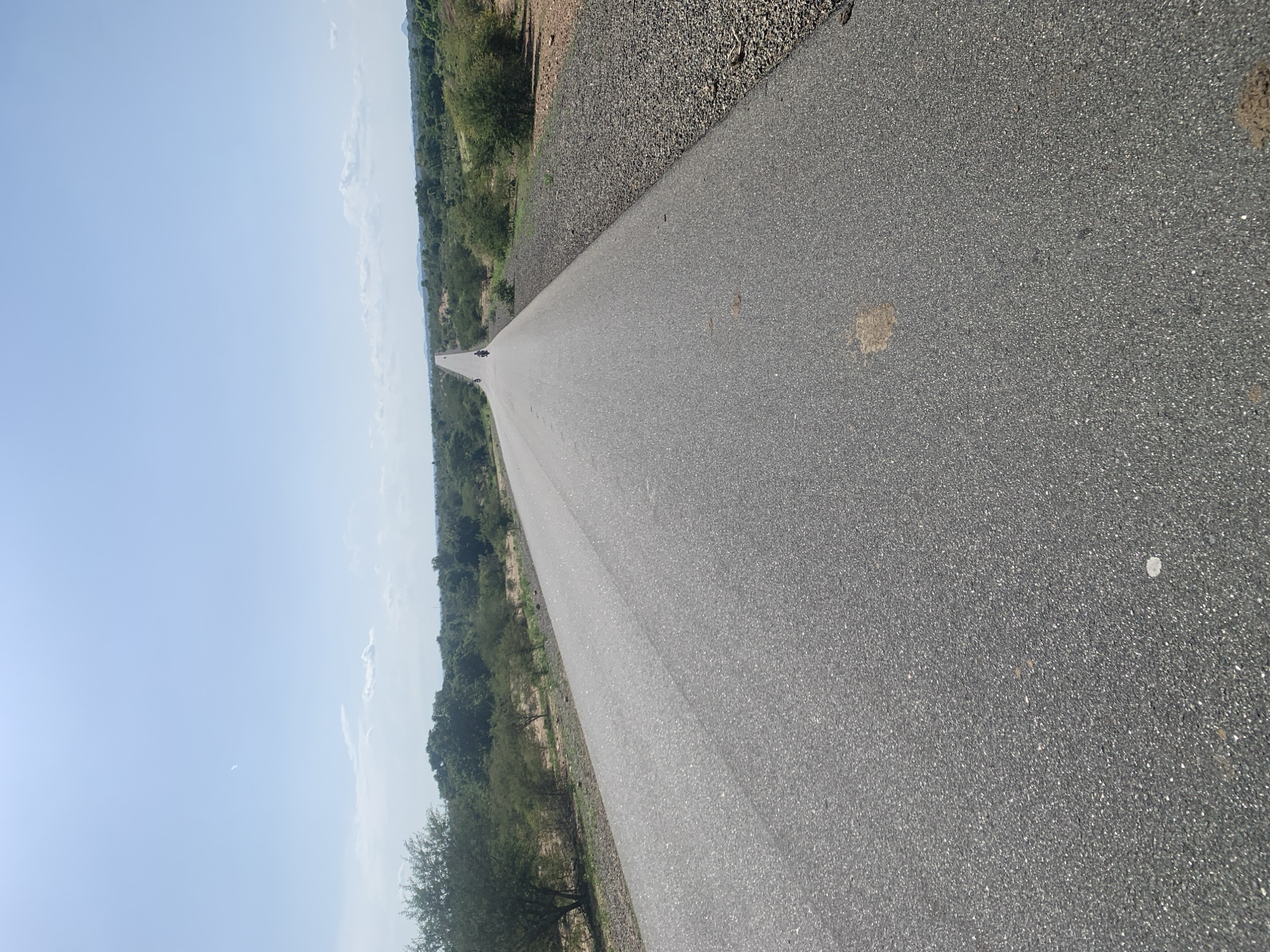 The image shows a straight, paved road stretching into the distance. The road is dark asphalt and appears to be in good condition. On either side of the road, there is dry, scrubby vegetation and scattered trees, suggesting a rural or semi-arid environment. To the left, a vast expanse of light blue sky dominates the frame, with a few wispy clouds visible. To the right of the road, there is a narrow strip of gravel or small stones.
In the distance, on the road, two figures on motorcycles are visible, heading away from the viewer. This indicates that the scene is likely taking place during daylight hours. The overall impression is one of a journey or travel through a somewhat desolate, yet open, landscape. The lack of prominent landmarks makes it difficult to pinpoint an exact location, but the environment is consistent with parts of Nigeria. There is no visible text in the image.
