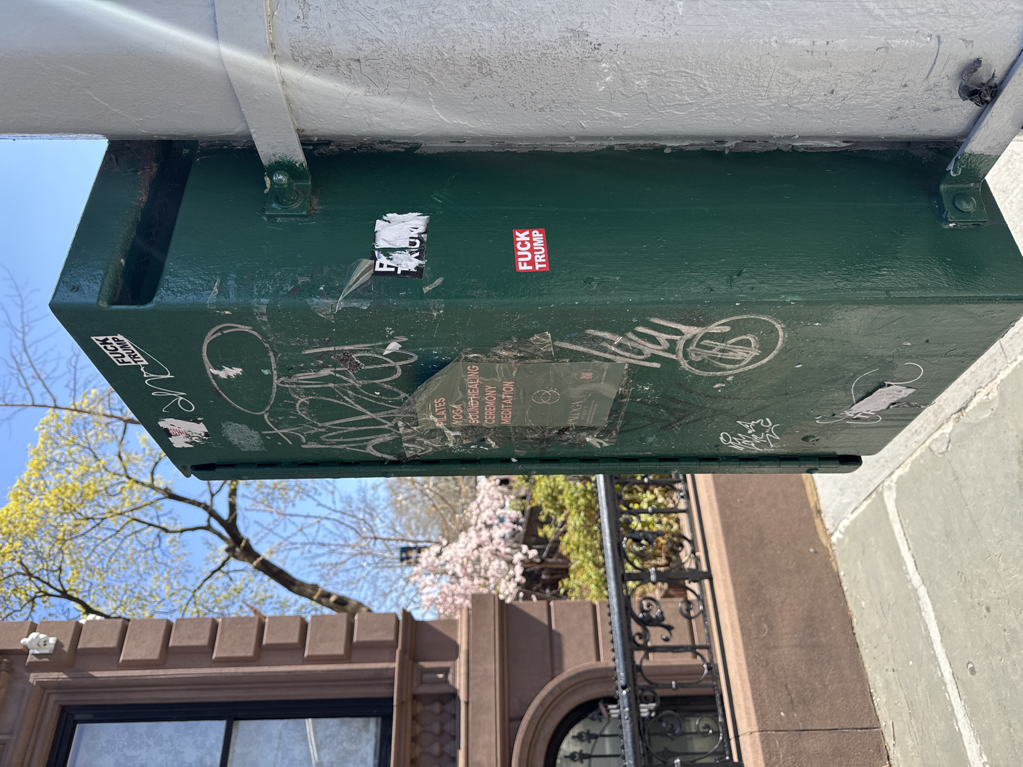The image captures a green metal box, likely a newspaper or postal box, affixed to a structure in New York City. The box is heavily marked with graffiti, stickers, and remnants of removed posters. The graffiti includes various tags, while stickers bearing the phrase "FUCK TRUMP" are visible. One sticker alludes to fitness and spirituality practices, mentioning Pilates, Yoga, Sound Healing, Ceremony, and Meditation. The box is situated in an urban setting, with an ornate wrought-iron fence, a building with a dark window, and budding trees visible in the background. The environment suggests a neighborhood, likely in New York City, based on the architectural style and street furniture.