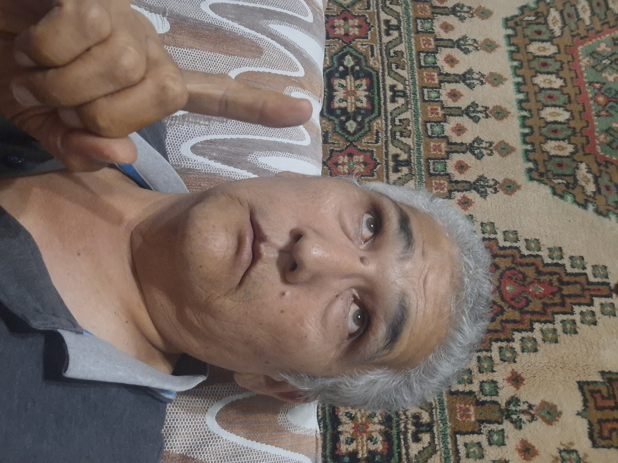 The image shows a close-up, self-taken photograph of an older man with gray hair and light brown skin. He is looking upwards and to his right. His left index finger is raised, and his mouth is slightly open. He is wearing a dark gray polo shirt with a light gray collar. The background appears to be a patterned carpet or tapestry with geometric designs in shades of brown, red, green, and white. There is also a section of white and brown patterned upholstery visible in the lower left portion of the image. The lighting suggests an indoor setting. There is no discernible information in the image to pinpoint its location to the city of Almaty, Kazakhstan.