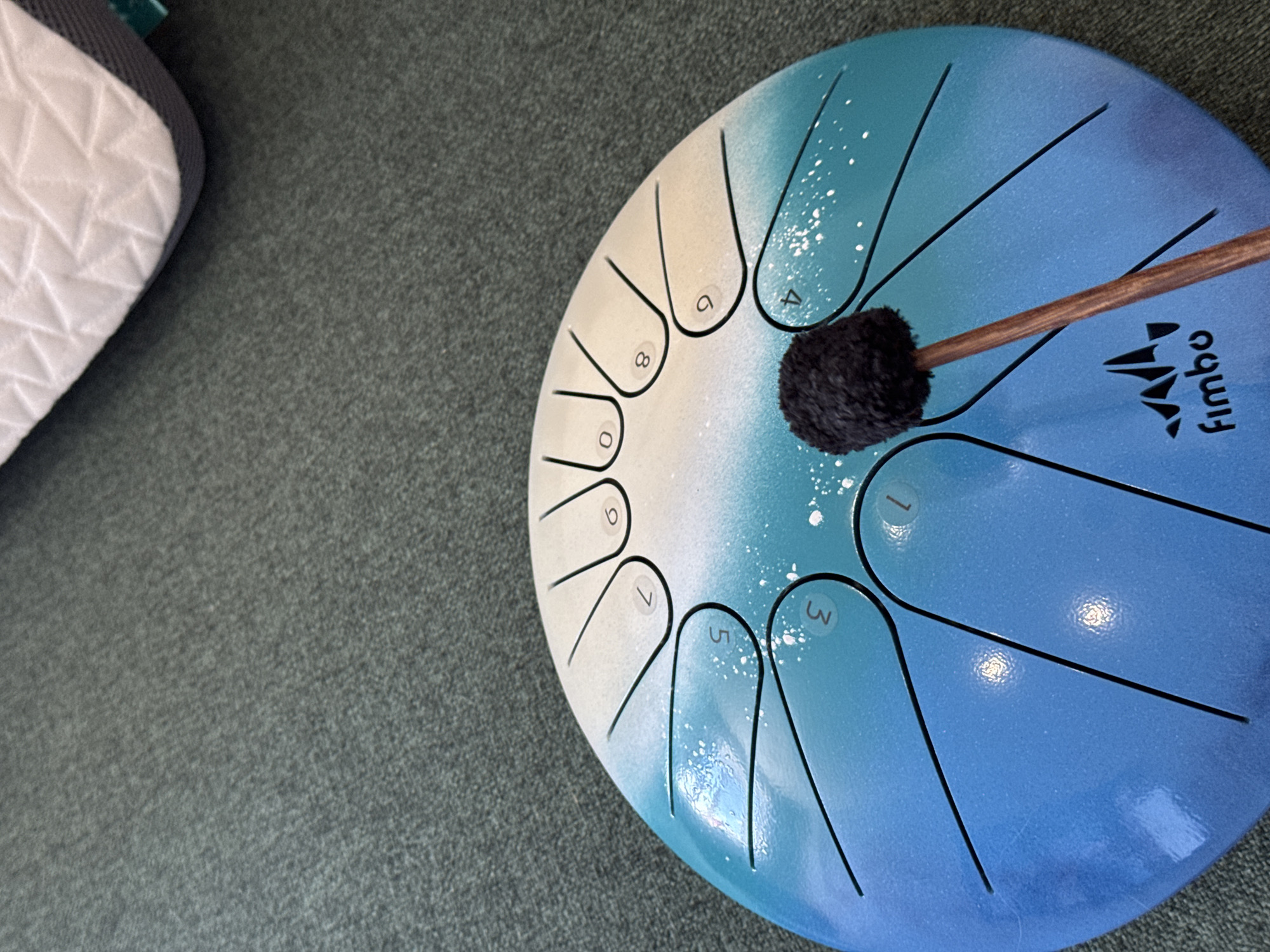 A top-down, medium shot captures a blue and white steel tongue drum resting on a textured, dark gray carpet. A wooden mallet with a fluffy black head is positioned to strike one of the drum's tongues. The drum is decorated with a gradient of blue to white, speckled with white dots that resemble stars. Black lines divide the drum into several sections, each marked with a white number, including 0, 3, 4, 5, 6, 7, 8, and 9. At the bottom of the drum, the word "Fimbo" is printed in a stylized font next to a triangular logo. A portion of a white and gray textured object, possibly a mattress or cushion, is visible in the upper right corner.