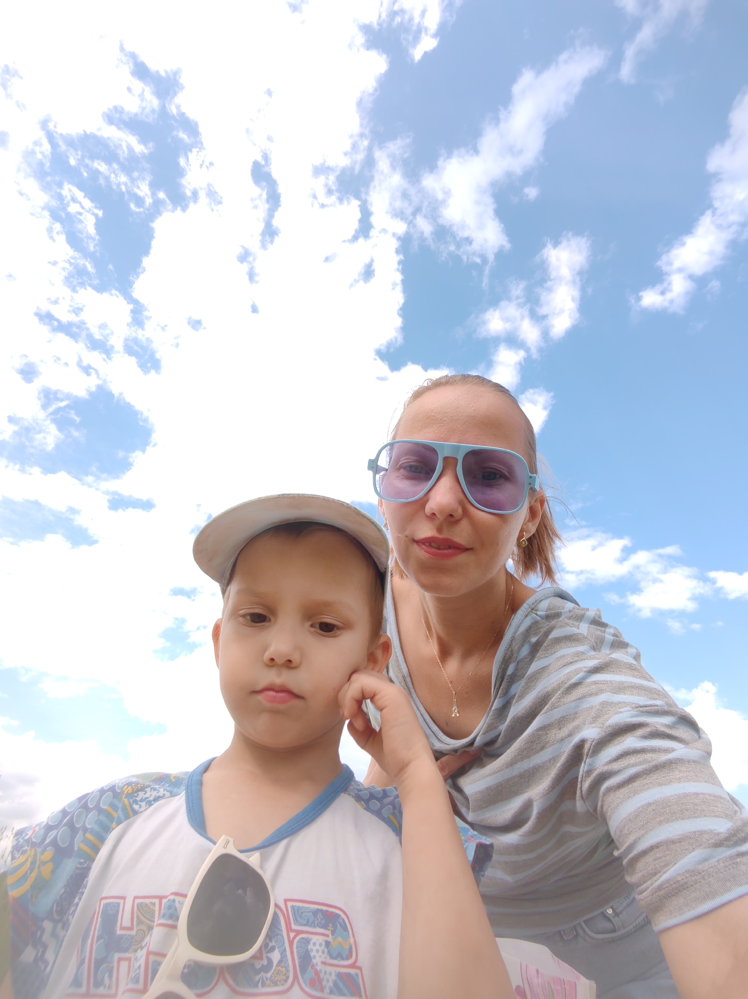 The image shows a selfie taken from a low angle, with the sky and clouds dominating the background. In the foreground, a young boy wearing a light-colored baseball cap and a white t-shirt with a blue neckline and a pattern on the sleeves is positioned on the left side of the frame. A pair of white sunglasses is hanging from the neckline of his shirt. To the right and slightly behind him, a woman is visible, wearing light blue tinted sunglasses and a gray and blue striped shirt. She has her left arm around the boy and her right hand on his shoulder. The scene appears to be outdoors, possibly in a park or recreational area, given the presence of a light blue sky with scattered white clouds. No specific landmarks or identifiable elements within the city of Чухлэм, Russia, are visible in the image.