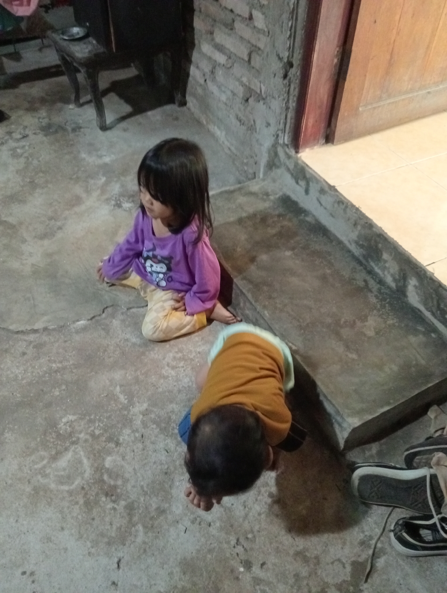 In Campago Barat, Indonesia, a young child wearing a purple shirt and light-colored pants sits on a textured concrete floor. The child has dark hair and is looking downwards. Nearby, another child, wearing an orange shirt over blue pants, is crawling on the floor, also looking downwards. The environment appears to be an outdoor or semi-outdoor space, with a low, grey concrete wall and a wooden door visible in the background. There is also a dark-colored wooden stool or table with a small plate on it in the upper left quadrant. Several pairs of dark-colored shoes are scattered on the floor to the right of the crawling child. The lighting suggests it may be evening or a dimly lit area.