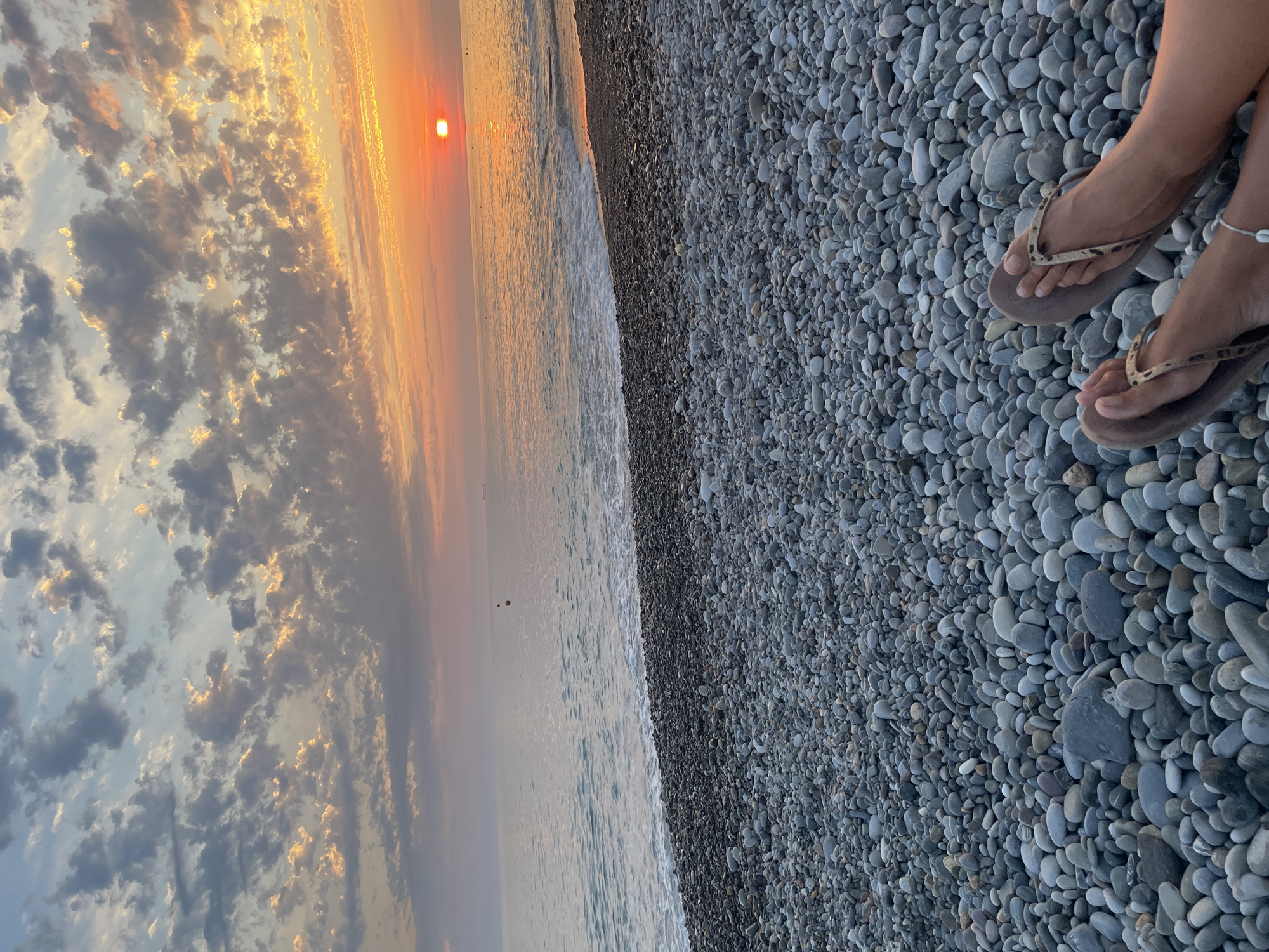 The image depicts a serene beach scene at sunset, with a focus on a person's feet in flip-flops resting on a pebble-covered shore. The sun, a bright orange orb, is setting over a calm ocean, casting a warm glow on the water and the scattered clouds above. The beach is composed of numerous smooth, grey pebbles. In the distance, a buoy and a ship are visible on the water. The overall atmosphere is one of tranquility and natural beauty.