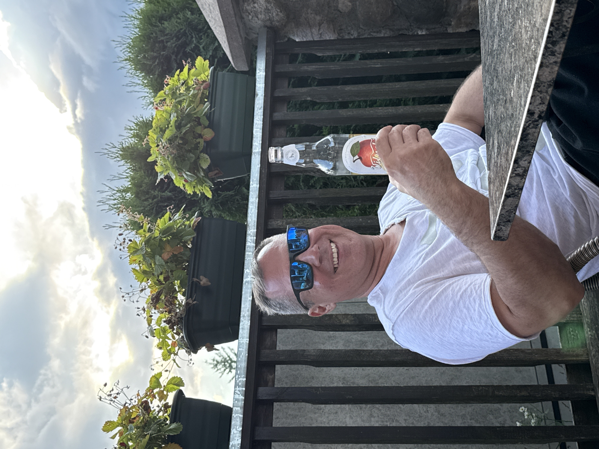 A man is holding a clear glass bottle with a light-colored liquid and a red apple label. He is wearing a white t-shirt and sunglasses with blue lenses. The man is smiling and looking at the camera. He is sitting at a table outdoors. In the background, there are potted plants and a wooden fence. The sky is cloudy. The photo was taken in Rūdiškės, Lithuania.
