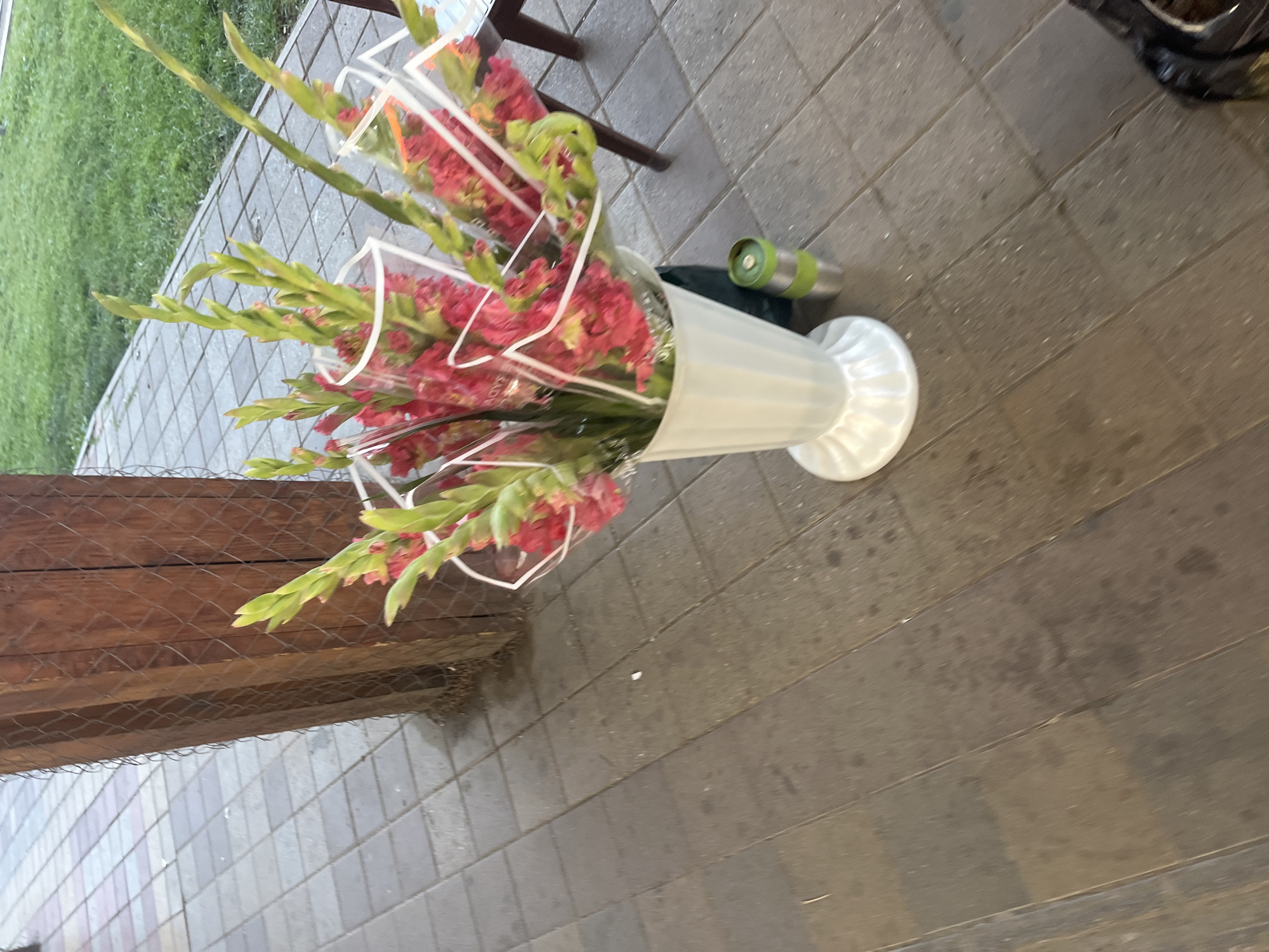 The image depicts a bouquet of gladiolus flowers in a white vase placed on a tiled walkway. The walkway is made of rectangular stone tiles. To the left of the walkway, there is a wooden structure with a mesh fence. Beyond the walkway, there is a grassy area. A metallic cup is visible next to the vase. The scene appears to be outdoors, possibly in a public space or a garden area. The image does not provide specific details to pinpoint a location within the city of Krasnodar, Russia. There are no people or actions visible in the image.