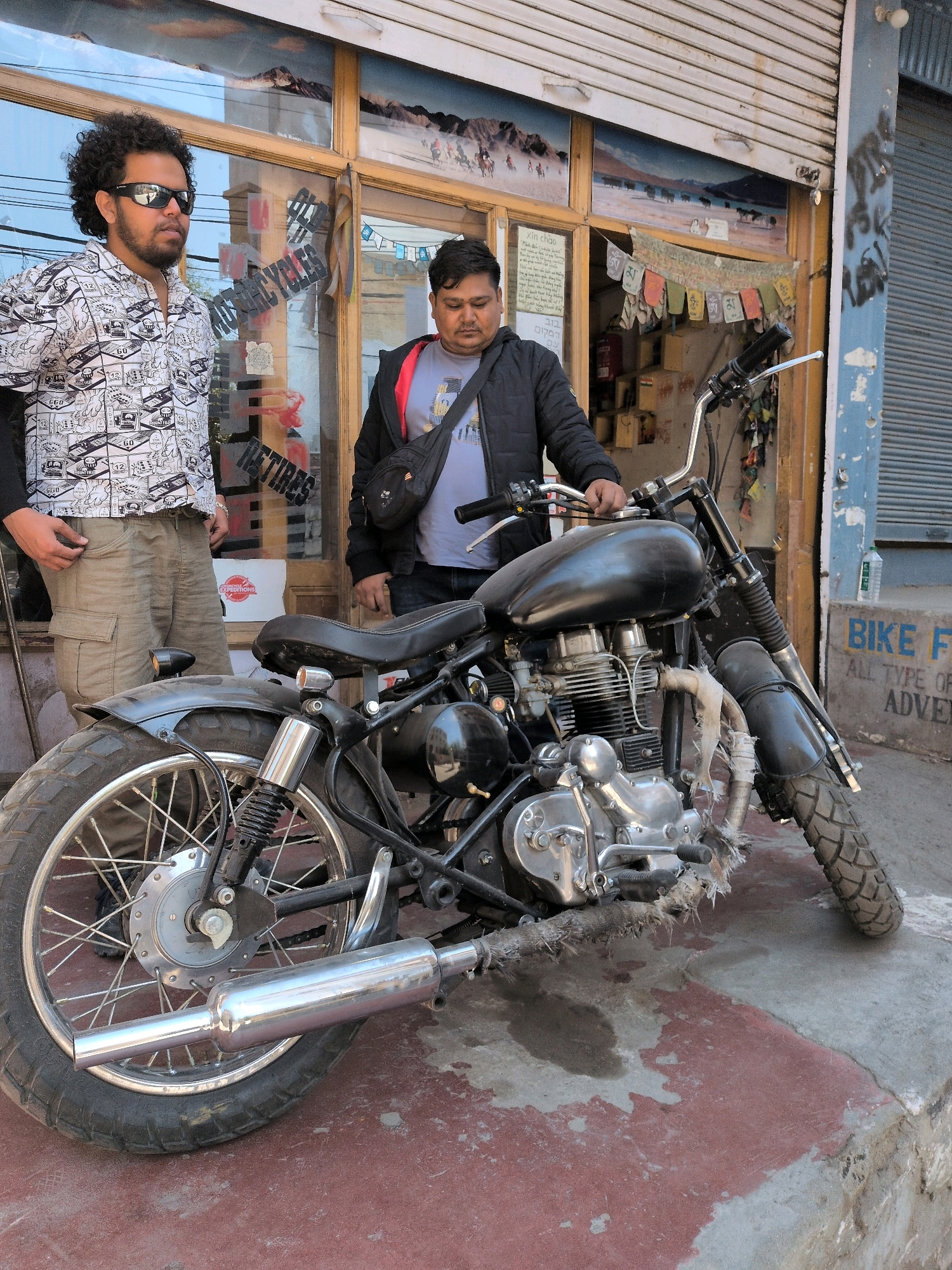 Two men stand next to a custom-built motorcycle in Leh, India. The man on the left, wearing sunglasses and a white patterned shirt with cargo pants, stands slightly forward, his left hand in his pocket. The man on the right, wearing a black jacket over a grey t-shirt and a black bag slung across his chest, stands behind the motorcycle, resting his right hand on the handlebars. The motorcycle, black and vintage-style, has a prominent exhaust pipe wrapped in a rough material and a custom seat. Behind the men, a shop with a large display window showcases what appears to be motorcycle parts and accessories. The window also displays colorful prayer flags and posters. The overall setting suggests a commercial establishment related to motorcycles in a street environment in Leh.