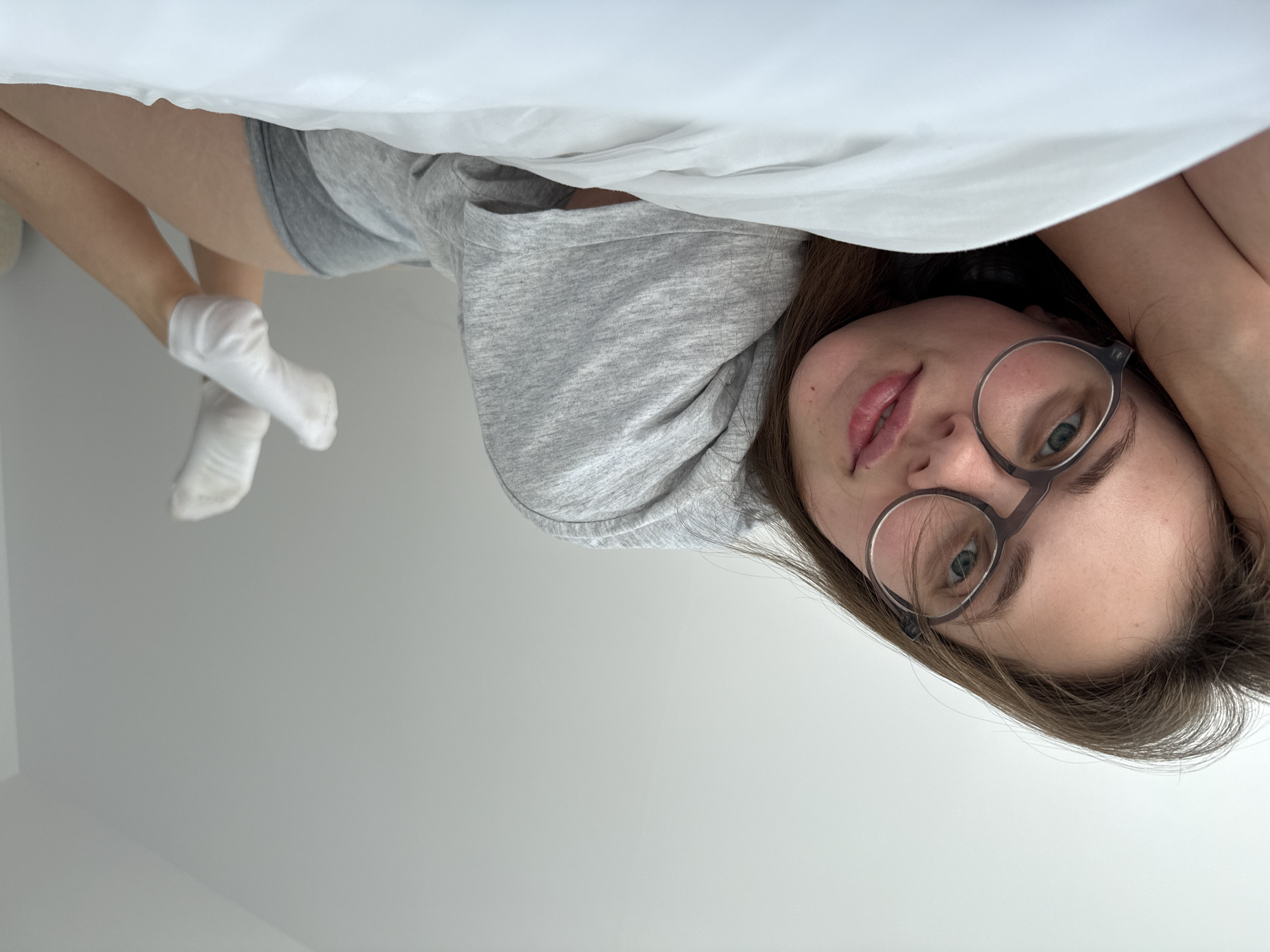 In Szklarska Poręba, Poland, a young woman with light brown hair and glasses is lying on a white bed. She is wearing a grey t-shirt and grey shorts, with white socks on her feet. Her head is resting on her arm, and she is looking at the camera with a slight smile. The background is a plain white wall.