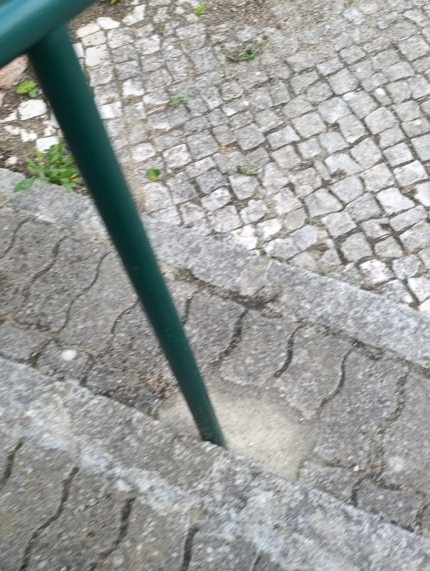 The image shows a green metal railing post embedded in a textured concrete surface. The railing post is dark green and extends vertically through the frame. The concrete surface appears to be part of an outdoor area, possibly a walkway or a staircase. The ground is paved with small, grey cobblestones arranged in a pattern. In the foreground, there is a section of concrete with a wavy, interlocking pattern. Small green plants are visible growing in some of the cracks between the paving stones. The lighting suggests it is daytime. The scene is likely located in an urban environment, given the paving and railing.