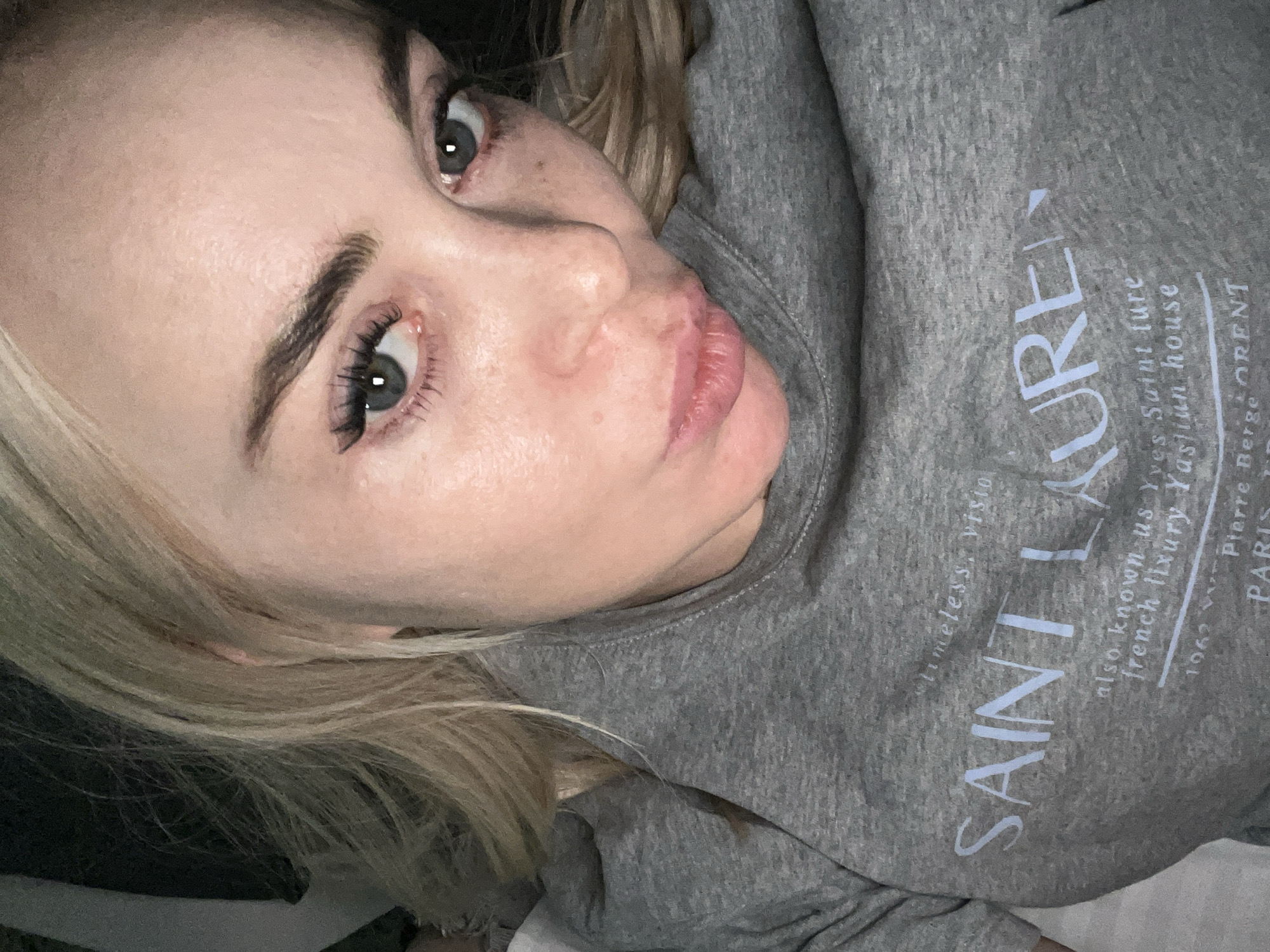 A close-up, high-angle, indoor selfie captures a woman with blonde hair and blue eyes. She is wearing a grey sweatshirt with text that appears to be related to "SAINT LAURENT." The background is dark and out of focus. The woman's head is tilted and she is looking towards the camera.