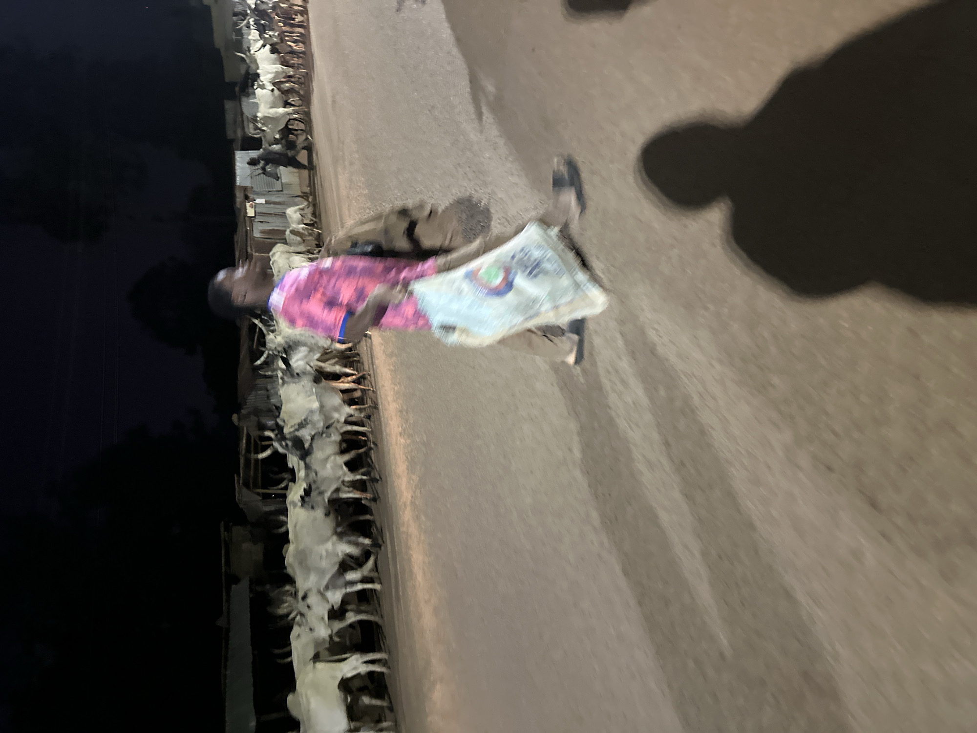 A man walks down a sidewalk in Bukuru, Nigeria, carrying a bag. The sidewalk is adjacent to a row of stalls or shops. It is nighttime, and the man's shadow is visible on the sidewalk. The stalls or shops appear to be dimly lit.