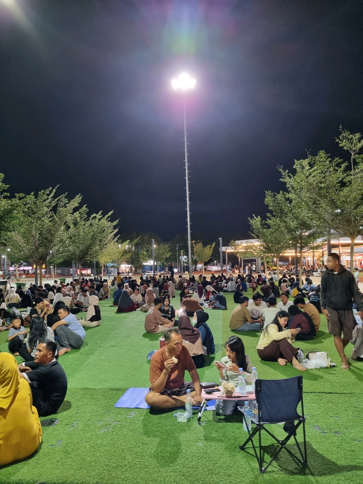 In Tanamodindi, Indonesia, a large gathering of people is seated on an artificial green turf at night. A tall light pole with bright floodlights illuminates the area. Many individuals are sitting in groups on the turf, some with food and drinks, suggesting a communal gathering or event. The atmosphere appears relaxed, with people interacting, looking at their phones, and eating. Trees line the background, and in the distance, buildings with illuminated interiors indicate a commercial or public space. The overall scene depicts a social gathering in an outdoor urban environment at night.
