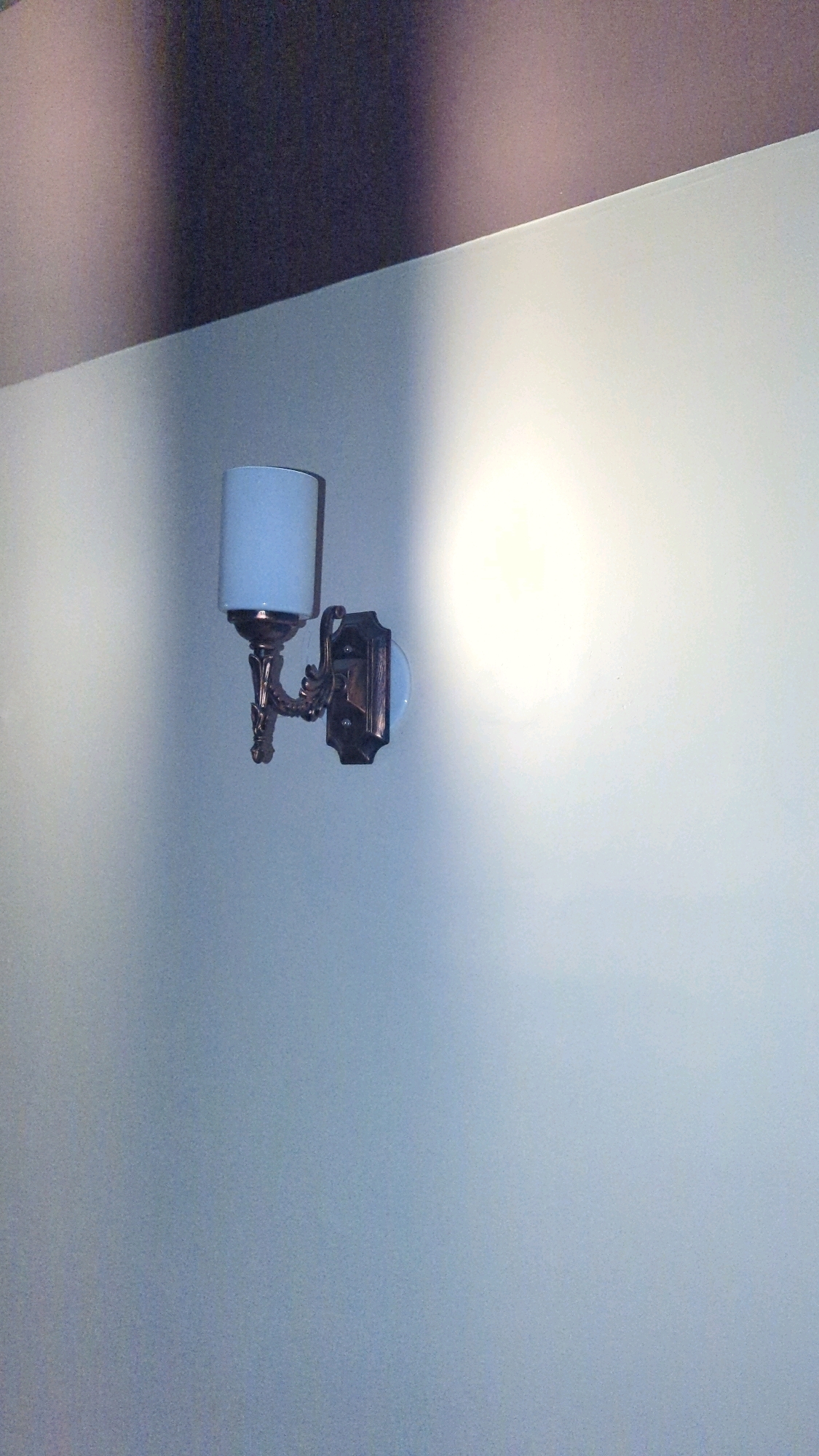 A wall-mounted light fixture with a blue cylindrical shade and an ornate bronze-colored metal base is depicted on a wall. The wall is painted a pale, cool tone, and a large, soft light source, possibly another light or a window, creates a bright glow on the wall to the right of the fixture. The upper portion of the image shows a darker, angled surface, possibly a ceiling or a shadow cast from above. The overall scene suggests an indoor environment. The image does not contain any specific visual cues to determine its location within Karachi Division, Pakistan.