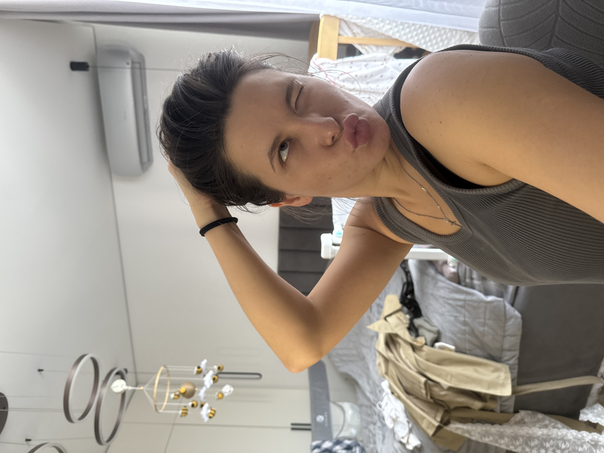 A woman is posing for a selfie. She is winking and making a kissing face. She is wearing a gray tank top. Her hair is dark and pulled back. She is touching her head with her right hand. A black bracelet is on her wrist. A light-colored coat is draped over a gray surface behind her. A modern chandelier is visible in the background. An air conditioning unit is mounted on the wall. Based on image analysis, the location is estimated to be a room within a residential building in Odintsovo, Russia.