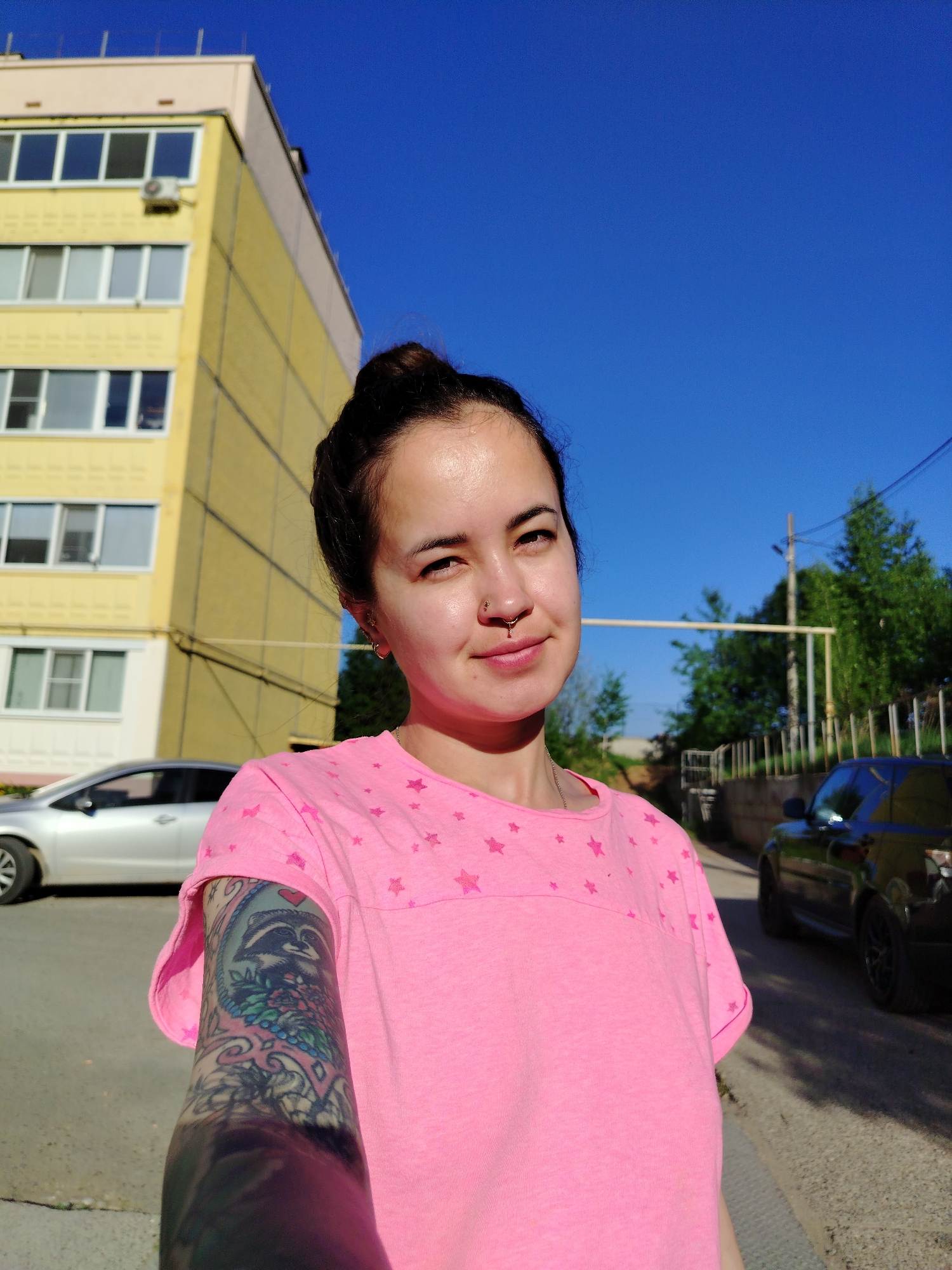 A young woman with dark hair pulled into a bun poses for a selfie in Noviy, Russia. She wears a pink t-shirt with a star pattern and has a nose ring. Her left arm is covered in a colorful tattoo. Behind her, a yellow apartment building stands to the left, and a black SUV is parked on the right. The sky is clear and blue. A silver sedan is also visible near the apartment building. Utility poles and trees are visible in the background. The scene is brightly lit, indicating it is daytime.