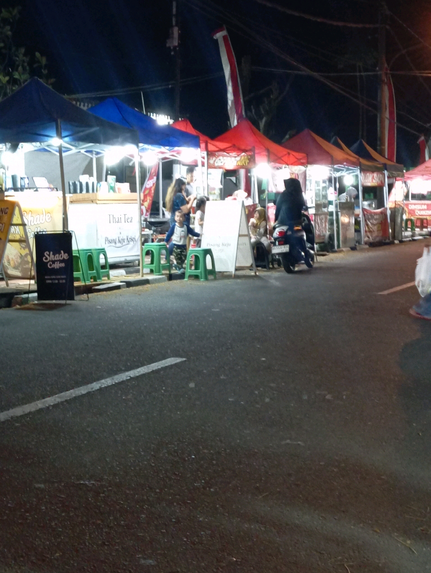 This is a nighttime, street-level, outdoor photograph of a food stall area, likely a night market, in the city of Magelang, Indonesia. The foreground shows an asphalt road with a white lane marking. In the midground, several food stalls are set up under colorful tents. One stall on the left has a sign that reads "Shade Coffee." Several green plastic stools are placed in front of the stalls.
A group of people are visible near the stalls. On the left, a child in a blue and white shirt is standing next to a woman in a dark blue top and a child in a white dress. Further to the right, a person in a black hijab is riding a dark-colored scooter, with another person, also in a hijab, seated behind them. Other figures are blurred in the background.
The stalls are illuminated by overhead lights, casting a warm glow. Some stalls have banners and displays, suggesting a variety of food and drink are being offered. The background is dark, indicating it is nighttime, with some electrical wires and poles visible overhead. The overall impression is a lively, albeit somewhat dimly lit, commercial street scene.