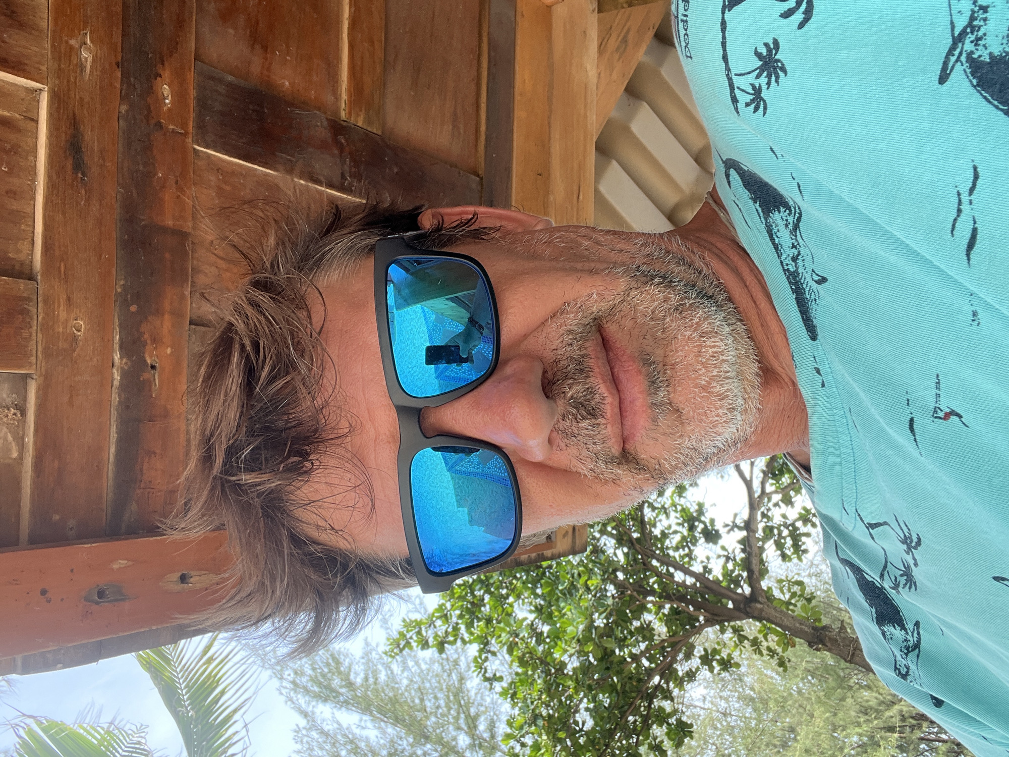 The image is a close-up, low-angle selfie of a man wearing sunglasses. He has tousled brown hair and a graying beard. The sunglasses have bright blue mirrored lenses that reflect a tropical environment, including a palm tree and a glimpse of the person taking the photo holding a phone. He is wearing a turquoise t-shirt with a black graphic print of birds and palm trees. Behind him, a wooden ceiling structure is visible, and to his left, lush green foliage and more palm trees are present, suggesting an outdoor setting in a tropical or subtropical climate. The overall impression is one of casual relaxation in a warm, sunny environment.