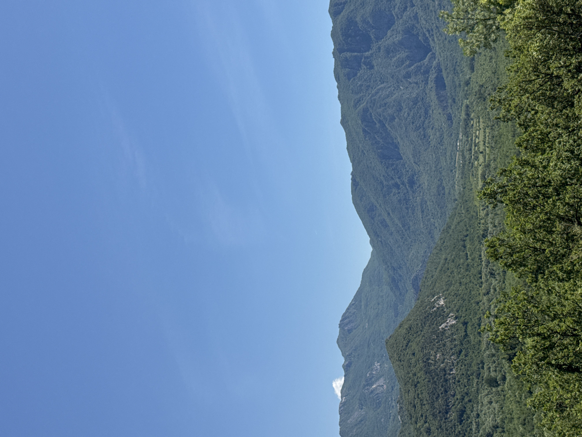 The image depicts a mountainside landscape under a clear, blue sky. Lush, green vegetation covers the slopes of the mountains, with trees visible on the right. The scene is likely located in a rural area. No people are visible. The location is in Sotonići, Montenegro.