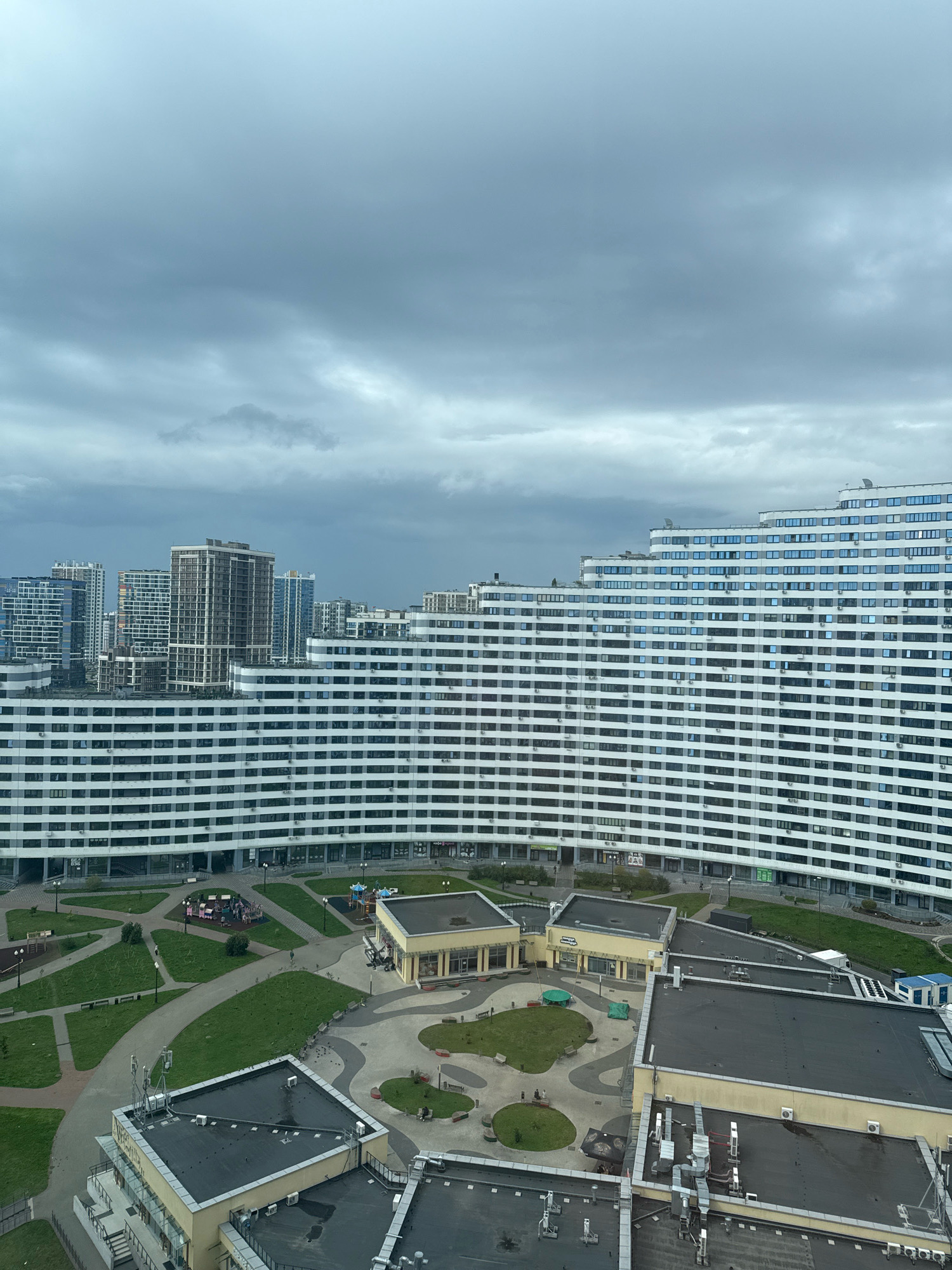 The image shows a panoramic view of the residential complex "Minsk World" in Minsk, Belarus. The complex is characterized by its modern architecture, with a large, curved white building dominating the foreground. This building features numerous windows and balconies, giving it a sleek and contemporary appearance. In the background, other tall buildings and skyscrapers can be seen, forming a dense urban landscape.
The ground level of the complex includes green spaces, playgrounds, and pathways, suggesting a well-planned residential area with amenities for its residents. The sky above is overcast and cloudy, indicating a possibly gloomy or rainy day. The overall impression is one of a large, modern city with extensive residential developments.