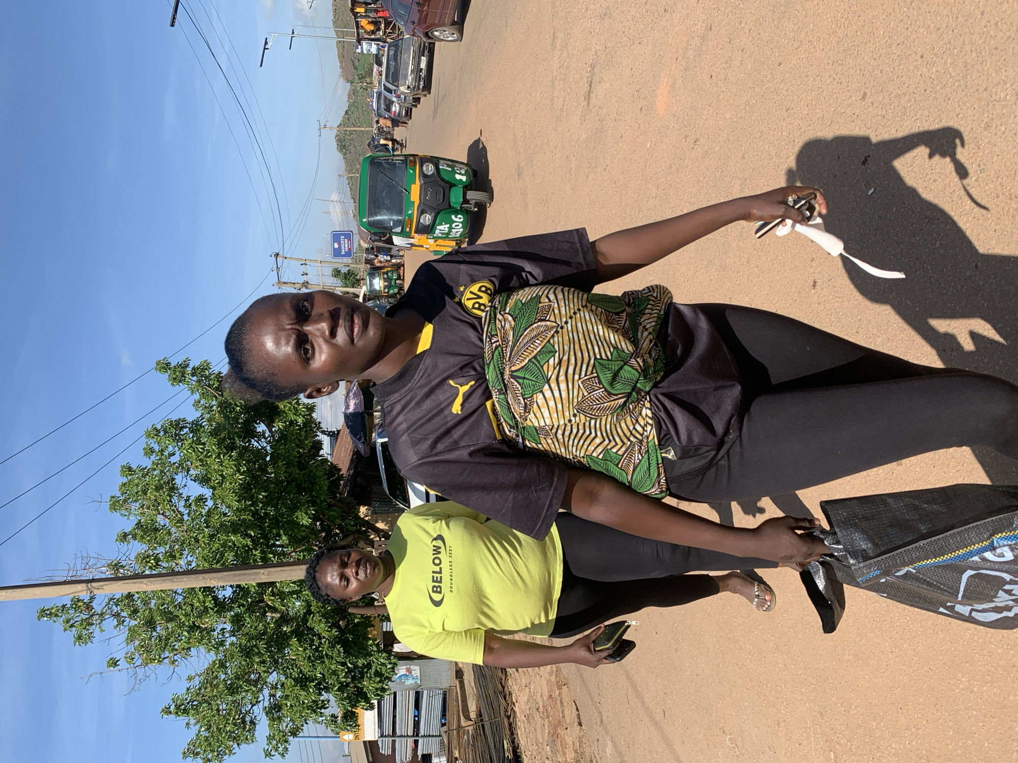 Two women walk down a dusty road in Gura Top, Nigeria. The woman in the foreground wears a black t-shirt with a yellow logo and a colorful patterned cloth tied around her waist. She carries a black bag in her left hand and a white object in her right. Behind her, a woman in a bright yellow t-shirt and black leggings walks, holding a black purse. The road is lined with shops and trees, and several vehicles, including a green auto-rickshaw, are visible. Power lines stretch across the clear blue sky.