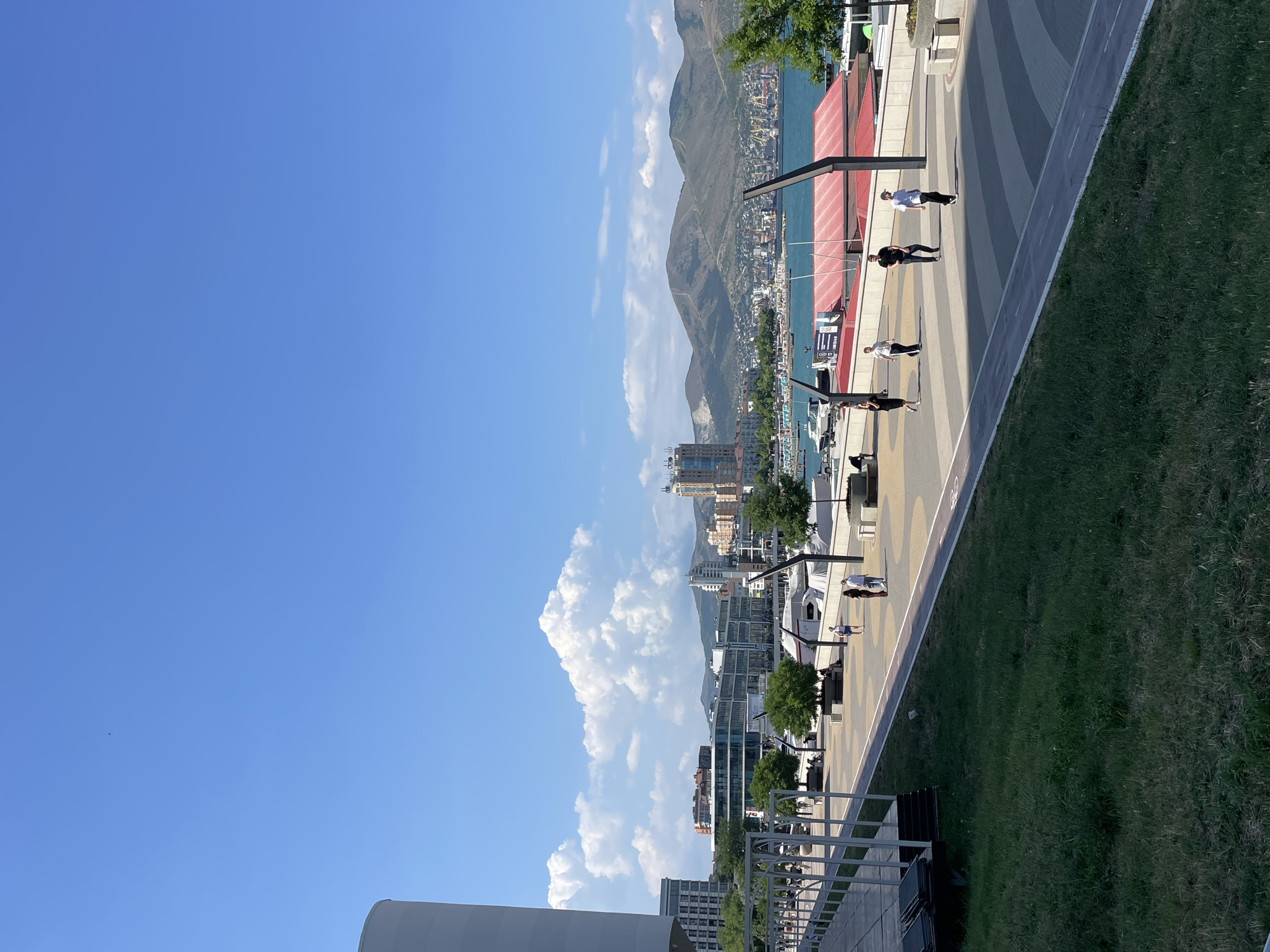 The image depicts a sunny day scene along the Novorossiysk embankment in Russia. In the foreground, a lush green lawn borders a paved walkway. The walkway, adorned with a geometric pattern of colored tiles, features a dedicated lane for cycling. Several individuals are seen strolling along the promenade. To the right, a body of water, likely the Black Sea, is visible with boats moored. Beyond the water, the cityscape of Novorossiysk unfolds, characterized by modern buildings, including high-rises, and a backdrop of rolling, green-covered mountains. A large industrial port with cranes is also discernible in the distance. The sky is a clear, vibrant blue, punctuated by fluffy white clouds.