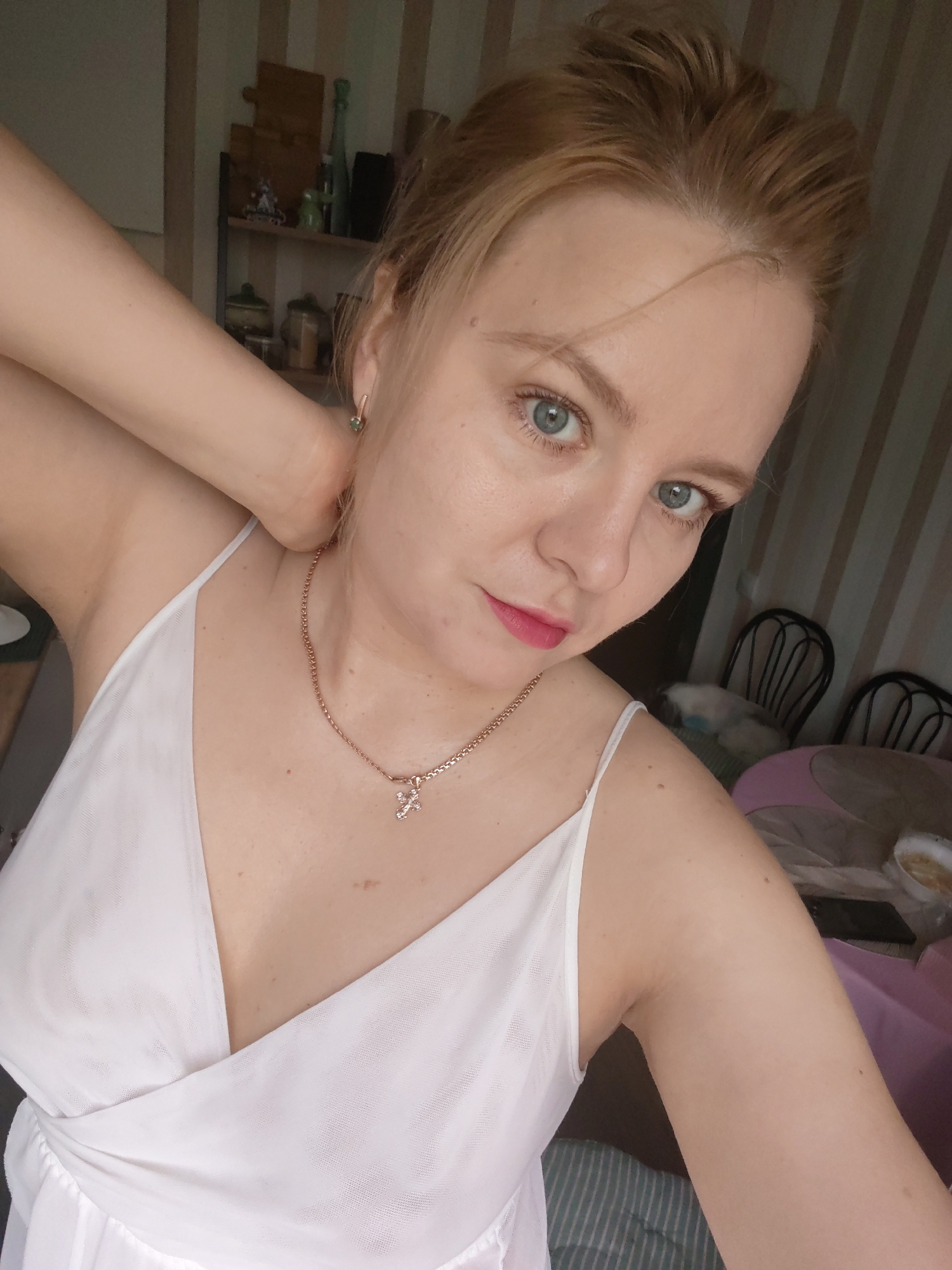 A woman with blonde hair and blue eyes, wearing a white camisole and a gold necklace with a cross pendant, poses for a selfie. Her left arm is raised, with her hand resting on the back of her neck. The background appears to be an indoor setting, possibly a room with shelves, chairs, and a table. The striped wallpaper suggests a home environment. There is no discernible information in the image that indicates a specific location within Izhevsk, Russia.