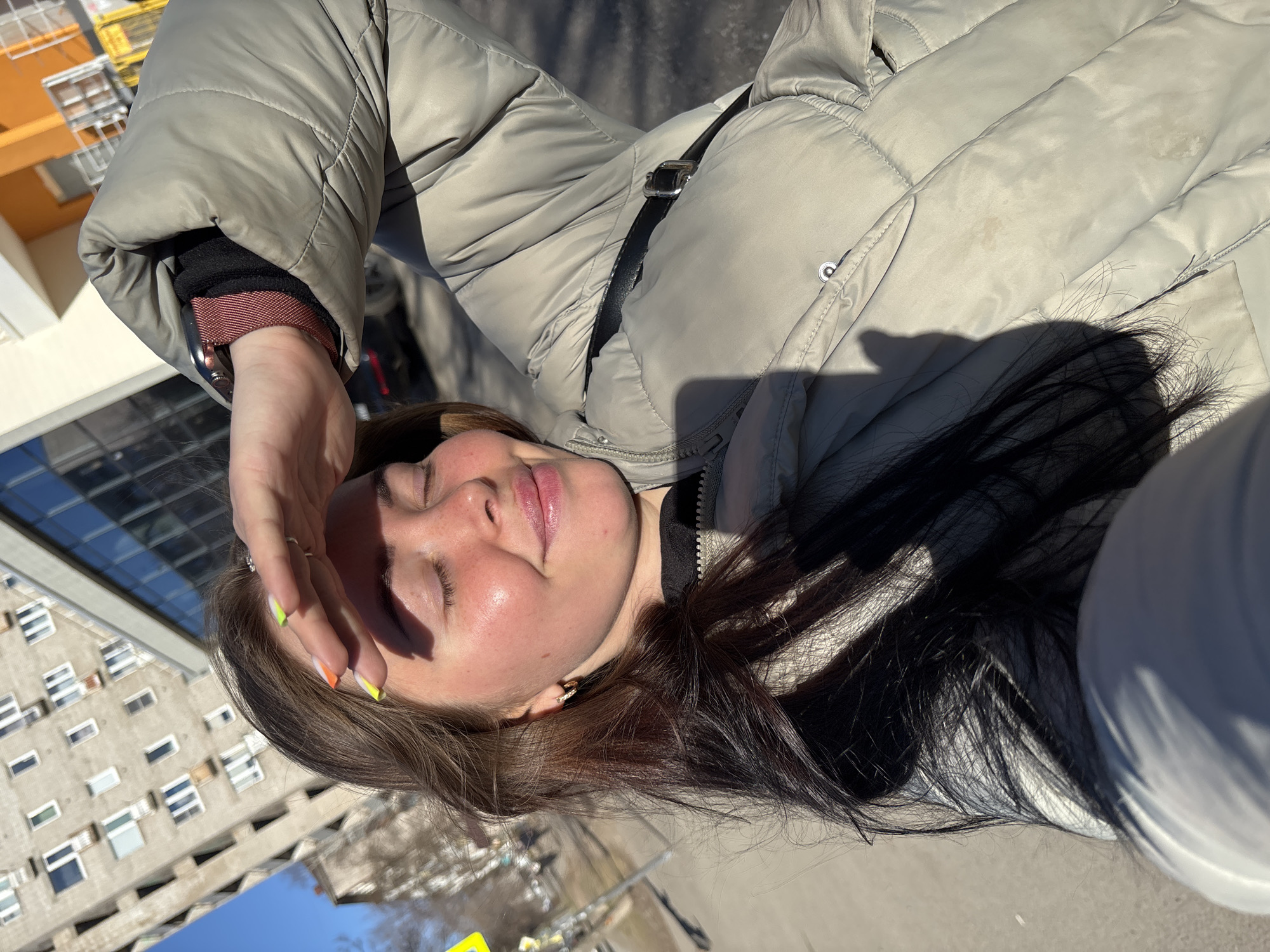 A young woman with brown hair and colorful nails is shielding her eyes from the sun with her hand. She is wearing a light gray puffer jacket. In the background is a view of urban architecture, including a building with multiple windows and a partially visible yellow and orange structure. The image was taken in Izhevsk, Russia.