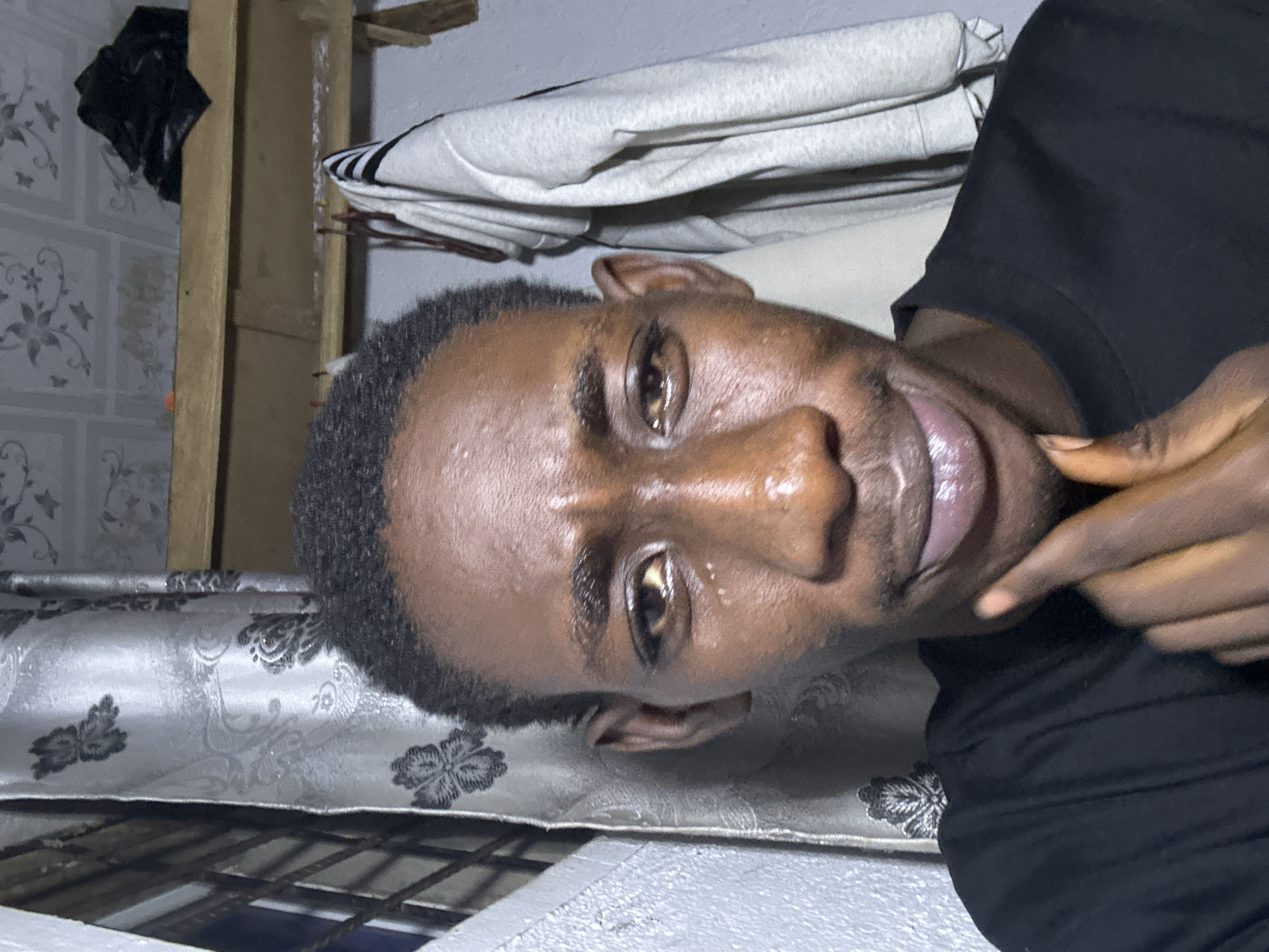 A medium shot captures a Black male posing for a selfie indoors. He has short, dark, curly hair and is wearing a black t-shirt. His right hand is raised to his chin, with his index finger touching his lips. The background consists of a patterned curtain on the left, a wooden shelf with items on it, and a grey wall with clothes hanging on the right. The lighting is dim, creating some glare on his skin, especially around his forehead and cheeks. The overall environment appears to be a bedroom or living space. The image does not provide any specific information to determine its location in Durumi, Nigeria.
