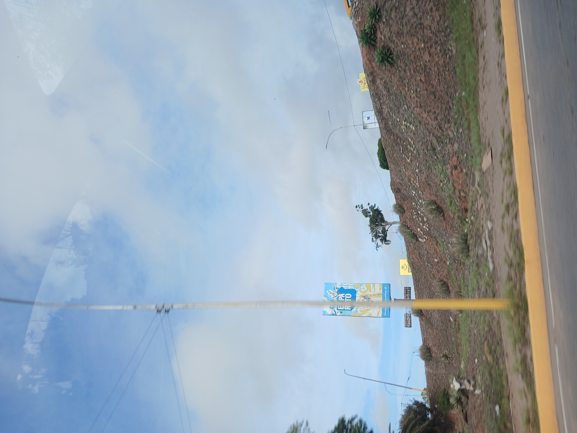This image captures a view of a hillside area in El Valle del Espíritu Santo, Venezuela, seen from the perspective of a moving vehicle. A yellow traffic barrier runs along the bottom edge of the frame. The hillside itself is covered in sparse vegetation, rocks, and some signs. Notably, there is a tall, slender yellow pole dominating the left side of the image, a billboard with blue and yellow graphics displaying text, and another smaller yellow sign further up the hill. The sky above is partly cloudy with visible reflections on what appears to be a window. The overall impression is a roadside view of a somewhat arid, developed landscape under a cloudy sky.