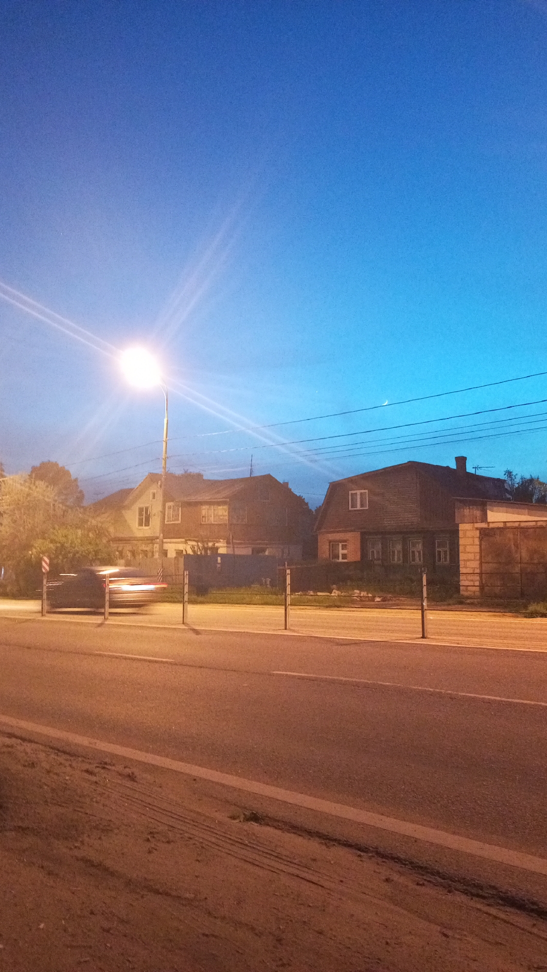 Captured at dusk in Юдино, Russia, the image reveals a road scene. A car, blurred in motion, is passing by a street lamp. Across the road are two houses with fences. Overhead are several power lines, and in the sky is a crescent moon. The overall impression is of a quiet residential neighborhood in twilight.