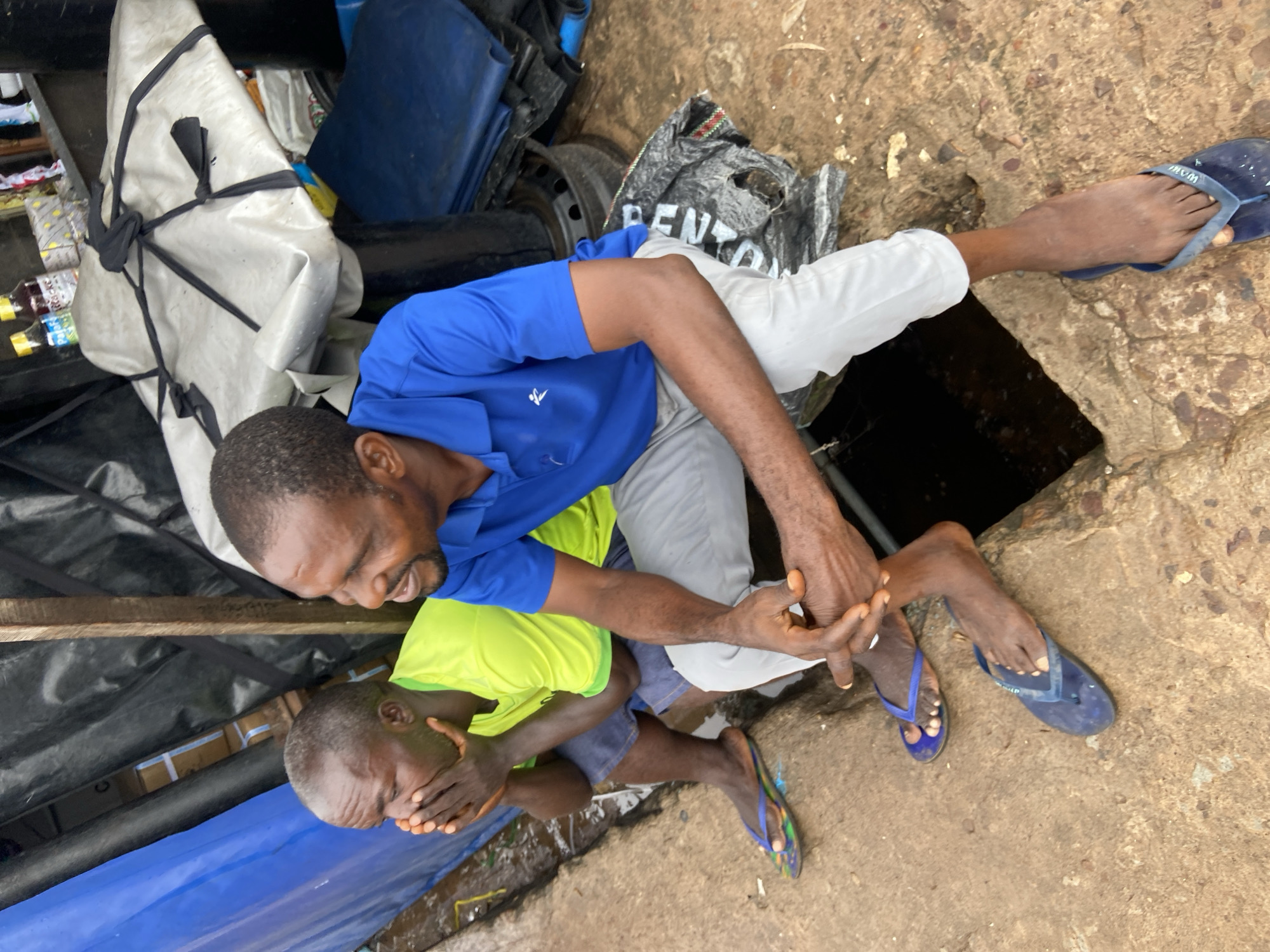 Two men, likely of Nigerian descent, are interacting outdoors in what appears to be an informal or somewhat cluttered setting. One man, wearing a blue shirt and light-colored pants, is kneeling or crouching, reaching towards the other man. The second man, younger and wearing a bright green shirt and shorts, is lying down and has his hands covering his face. His body is oriented in a way that suggests he might be distressed or in pain.
The ground is a rough, concrete-like surface, with some dirt and debris visible. There are various objects in the background, including what looks like tarpaulins, bundles wrapped in white material, a blue plastic container, and a discarded tire. A black plastic bag with white lettering is also present. The lighting suggests it might be daytime, but the overall scene appears somewhat dim or overcast. The presence of a visible opening in the ground, possibly a well or drain, is a significant detail in the setting. The man in the blue shirt is wearing dark flip-flops, and the younger man is wearing patterned flip-flops. The scene appears to be in Ogbunike, Nigeria. The overall emotion conveyed is one of concern or distress, particularly from the younger man's posture.