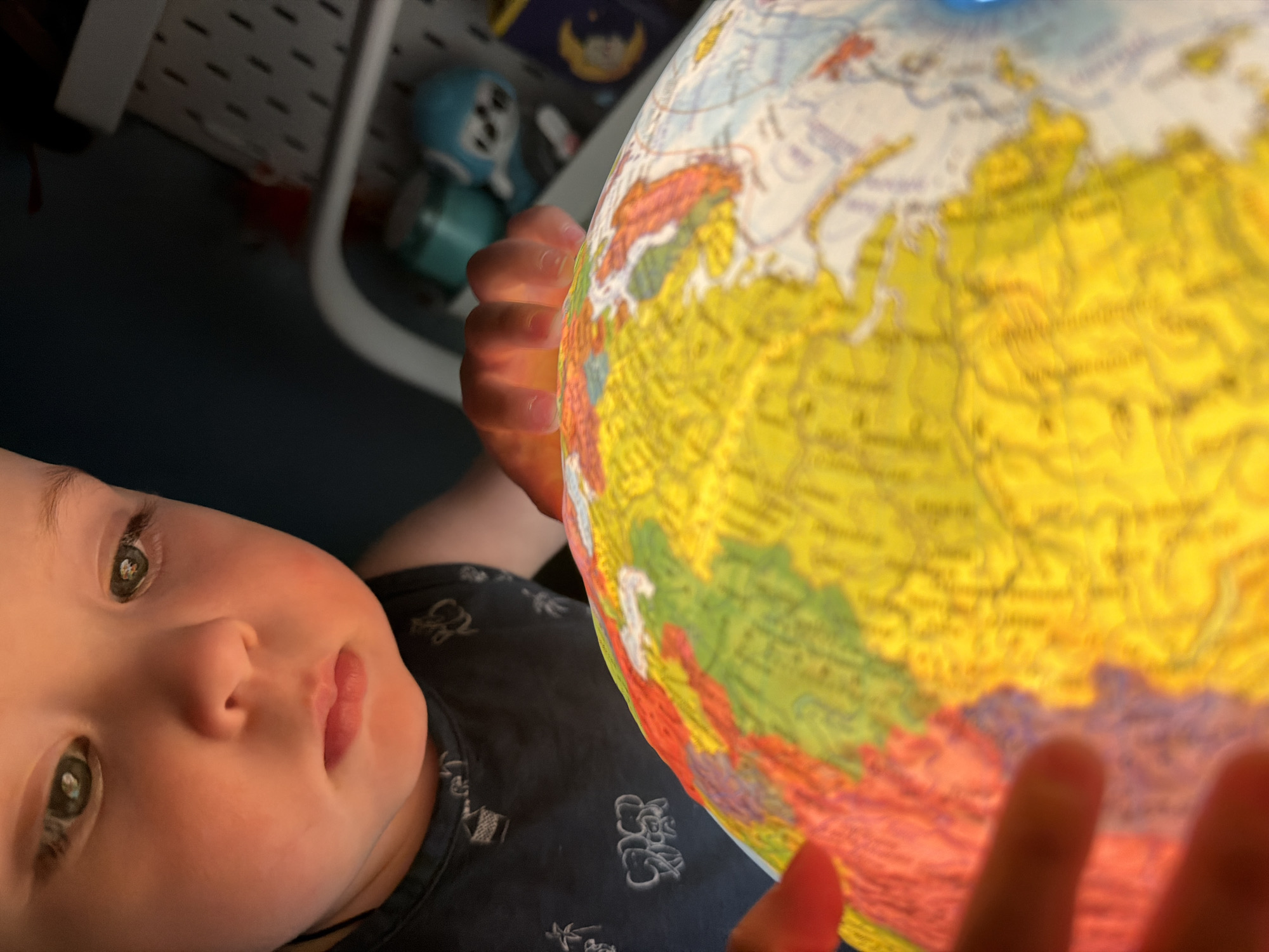 A close-up, low-angle shot shows a baby's face and hands interacting with a brightly lit globe. The baby, with light skin and wide, green eyes, looks curiously at the globe. They are wearing a dark blue t-shirt with a pattern of light-colored Asian-style figures. The baby's hands are visible, one on each side of the globe, holding it. The globe is illuminated from within, casting a warm glow and making the map details vibrant. The visible portion of the globe appears to be Eurasia, with yellow and red landmasses and blue oceans. The background is dark and out of focus, with some hints of shelves and objects, including what appears to be a blue toy and some paperwork. The lighting suggests an indoor setting, possibly a child's room or play area, and the mood is one of exploration and wonder. There is no visible text in the image.