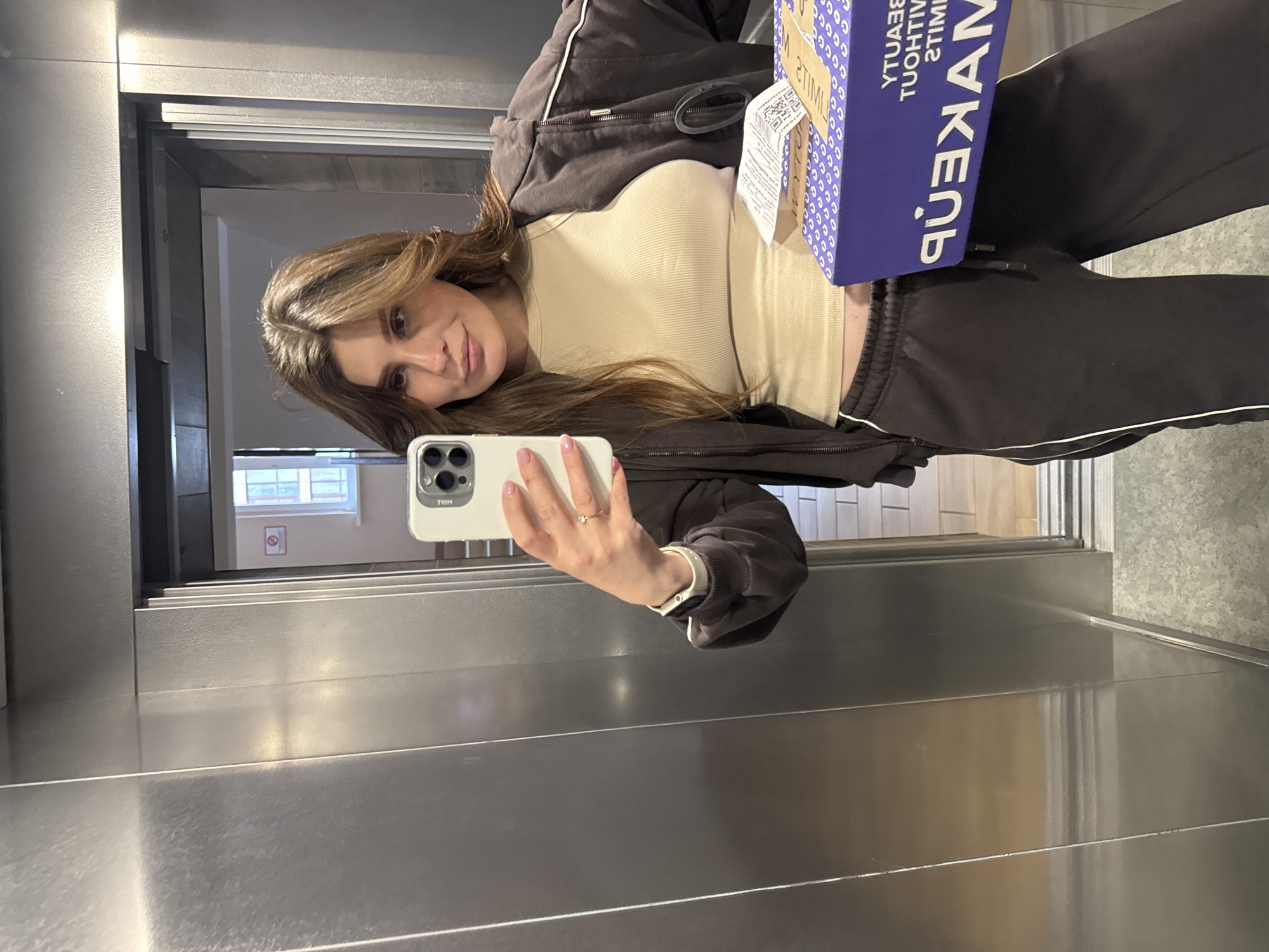 A young woman captures a mirror selfie in what appears to be an elevator or a space with highly reflective metallic walls, located in Sofiivska Borshchahivka, Ukraine. She has long, light brown hair and is wearing a beige ribbed top layered under a dark brown or black zip-up hoodie and matching tracksuit bottoms with white side stripes. She wears a ring on her right hand, which holds a light-colored iPhone with multiple camera lenses, its case subtly displaying "TRH". In her left hand, she holds a blue package. The package has white text that, when read in reverse due to the mirror reflection, says "MAKEUP" and "BEAUTY WITHOUT LIMITS", along with a QR code and a shipping label.
The reflections on the metallic walls show an indoor environment with a window and what seems to be a fire safety sign (a red circle with a diagonal line through an icon) on a light-colored wall. The woman looks directly at the camera with a neutral to slightly smiling expression. The lighting suggests daytime, possibly from the reflected window. The overall scene conveys a casual, everyday moment of someone receiving and documenting a package delivery.