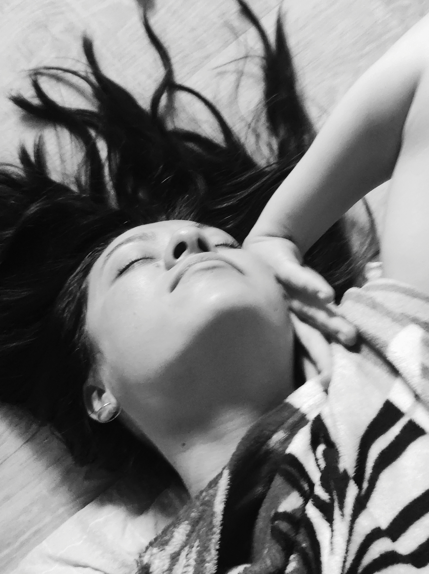 A grayscale medium shot depicts a person reclining with their head tilted back and eyes closed. Long, dark hair fans out around their head, contrasted against a light-colored surface with horizontal lines, likely a wooden floor. A child's hand rests on the person's face. The person is wearing a patterned garment, possibly a robe or blanket with dark zebra-like stripes on a light background. An earring is visible in the ear. The scene appears to be indoors, with no indication of a specific location within Unknown, Unknown.