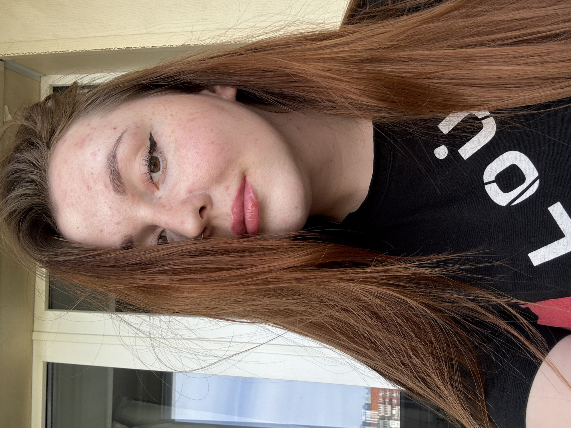 A portrait of a fair-skinned woman with reddish-brown hair is captured indoors, near a window. Her face is slightly tilted, revealing a light makeup application including winged eyeliner and lip gloss. She is wearing a black t-shirt with white lettering, part of which is visible. The window offers a glimpse of the exterior, featuring buildings identifiable as being in Izhevsk, Russia.