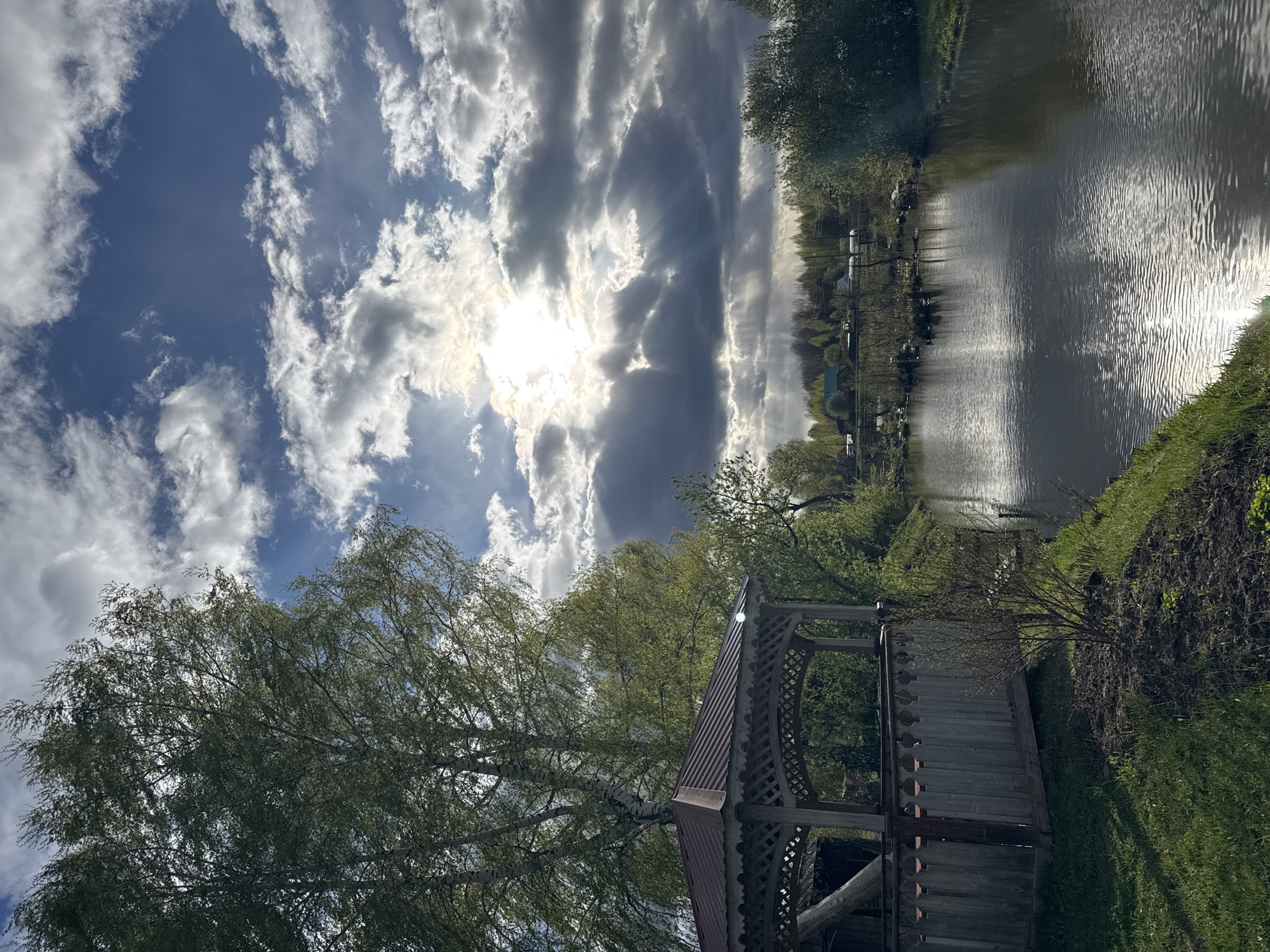 The image depicts a gazebo located near a body of water, possibly a lake or river, in Krilovo, Russia. The sky is partly cloudy with the sun visible behind the clouds. The gazebo is constructed of wood, with a decorative lattice design. A large tree with light green foliage is located adjacent to the gazebo. The body of water reflects the sunlight and surrounding trees. The shoreline is grassy and slightly elevated, with some bare soil patches. In the distance, buildings and trees can be seen on the opposite side of the water.