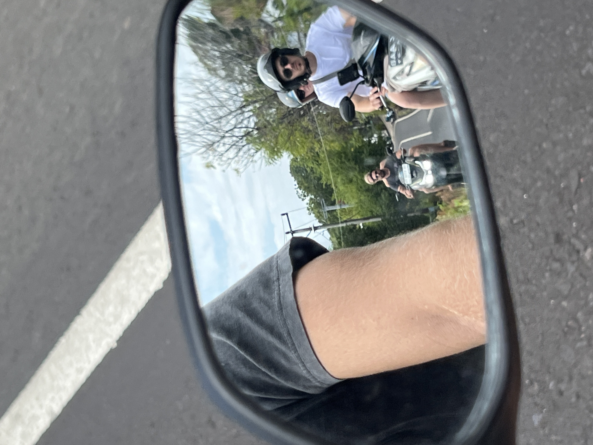 In the city of Abang, Indonesia, a reflection in a motorcycle's side mirror shows three individuals riding scooters on a road. The primary reflection features a person wearing a white t-shirt and sunglasses, with another person behind them. To the left, a third individual on a scooter is visible. The road surface is asphalt with a white painted line. Green foliage and trees line the sides of the road, with a cloudy sky overhead. The foreground of the image shows the arm and shoulder of the person taking the photo, wearing a dark t-shirt.