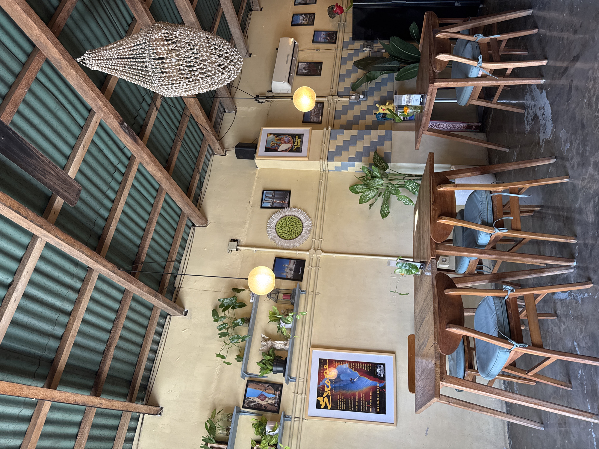 This image depicts the interior of a cafe in Kuta Selatan, Indonesia, likely a restaurant or a similar establishment given the dining tables and chairs. The space has a rustic and bohemian aesthetic. The ceiling is constructed with exposed wooden beams and corrugated green roofing, allowing natural light to filter through. A large, ornate chandelier made of white beads hangs from the ceiling. The walls are painted a pale yellow, with a decorative tiled section featuring a diamond pattern of blue and beige squares lower down.
Several wooden dining tables and chairs with light blue cushioned seats are arranged in the dining area. The tables are simple, wooden designs with angled legs.
The walls are decorated with various framed pictures and posters, as well as several potted plants on shelves. One prominent framed poster on the left wall features a graphic design with text. To the right, near the corner, is an air conditioning unit mounted on the wall.
The floor appears to be concrete with some water stains or discoloration. In the background, a large black appliance, possibly a refrigerator, is visible. A small yellow figurine of the character Pikachu is placed on a shelf near the tiled wall section, adding a playful touch to the decor. The overall impression is of a casual, artsy, and comfortable dining environment.