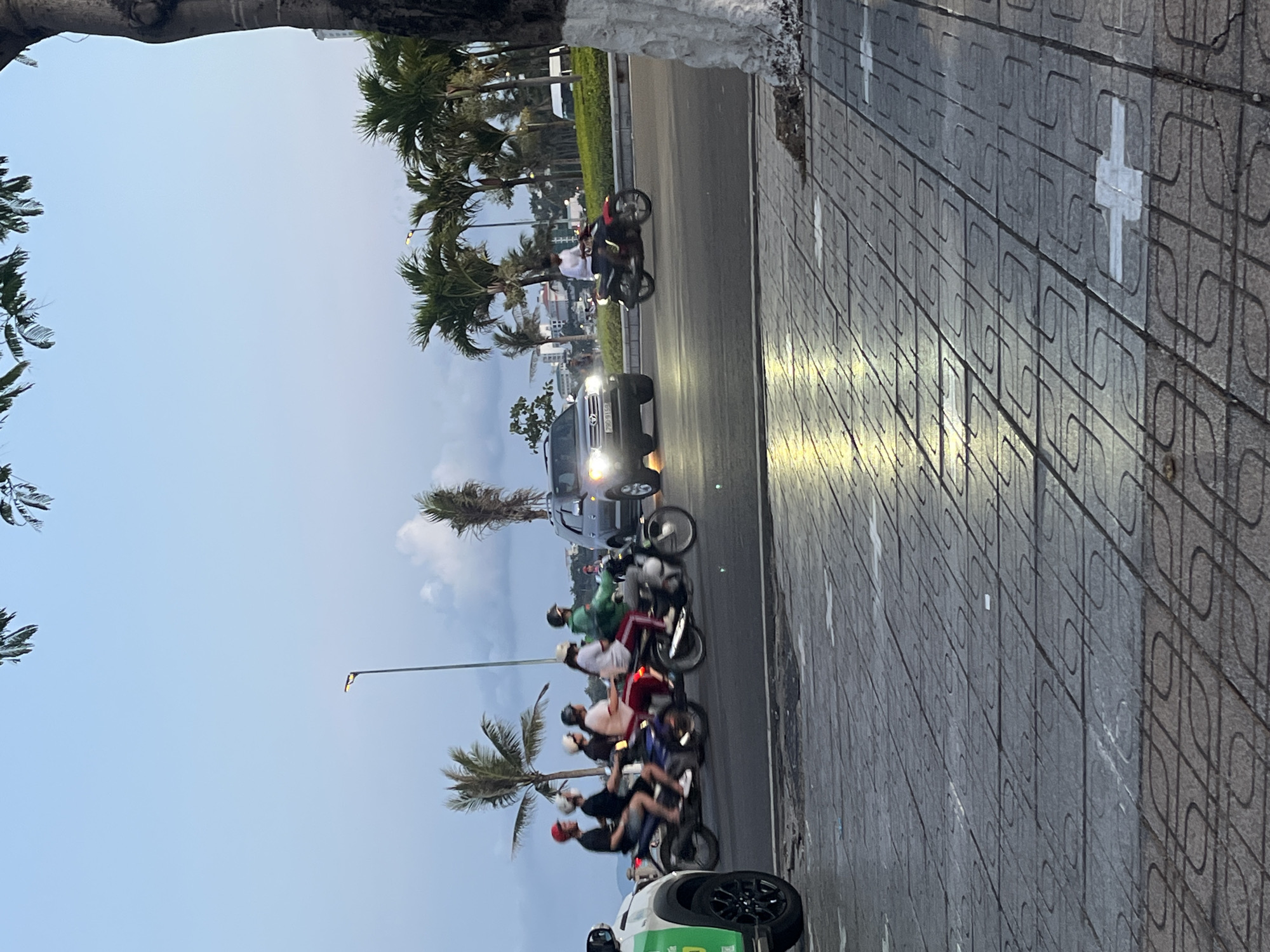 The image depicts a street scene in Bac Nha Trang Ward, Vietnam. A line of motorbikes carrying passengers are traveling on the road. A silver SUV with illuminated headlights is also present. Palm trees line the roadside. The sidewalk is paved with patterned gray tiles marked with white crosses. The sky is overcast.