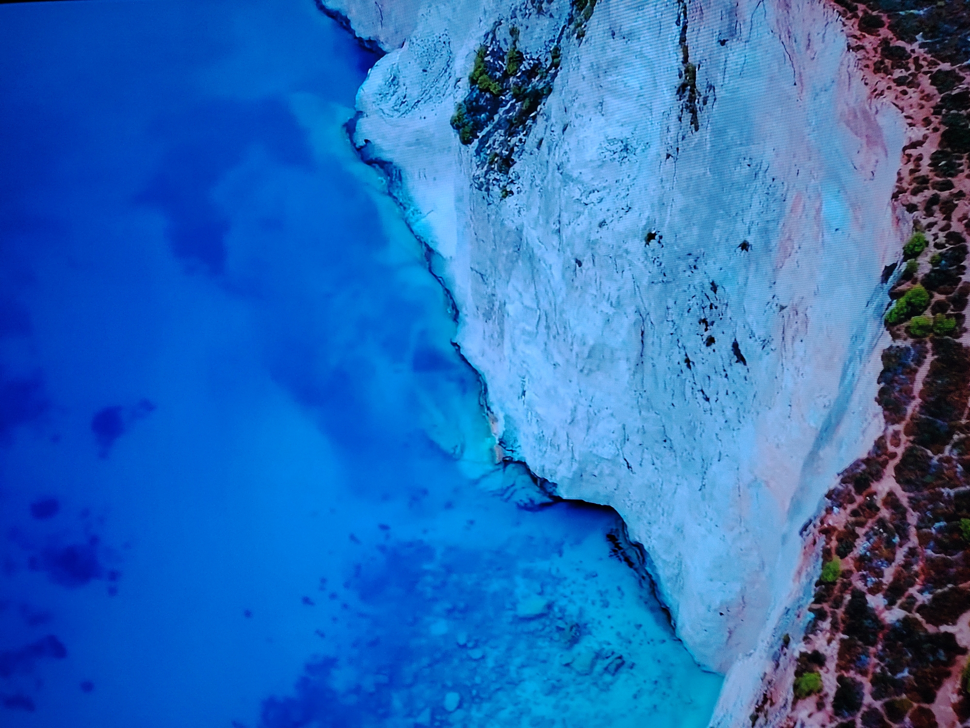 This is an aerial, high-angle, eye-level, full shot of a coastal cliff face meeting the ocean. The cliff is composed of pale, layered rock, with patches of green vegetation growing on top and interspersed in crevices. The ocean is a vibrant, deep blue, with lighter turquoise areas indicating shallows or underwater features. The water is calm, with some subtle patterns suggesting currents or underwater topography. The top of the cliff transitions into a rugged, earthy terrain covered in dense, low-lying vegetation with reddish-brown and green hues. The scene is natural and devoid of any human presence or man-made structures. The location is not identifiable as being within the city of Huntingdonshire, United Kingdom, as the image depicts a natural coastal landscape.