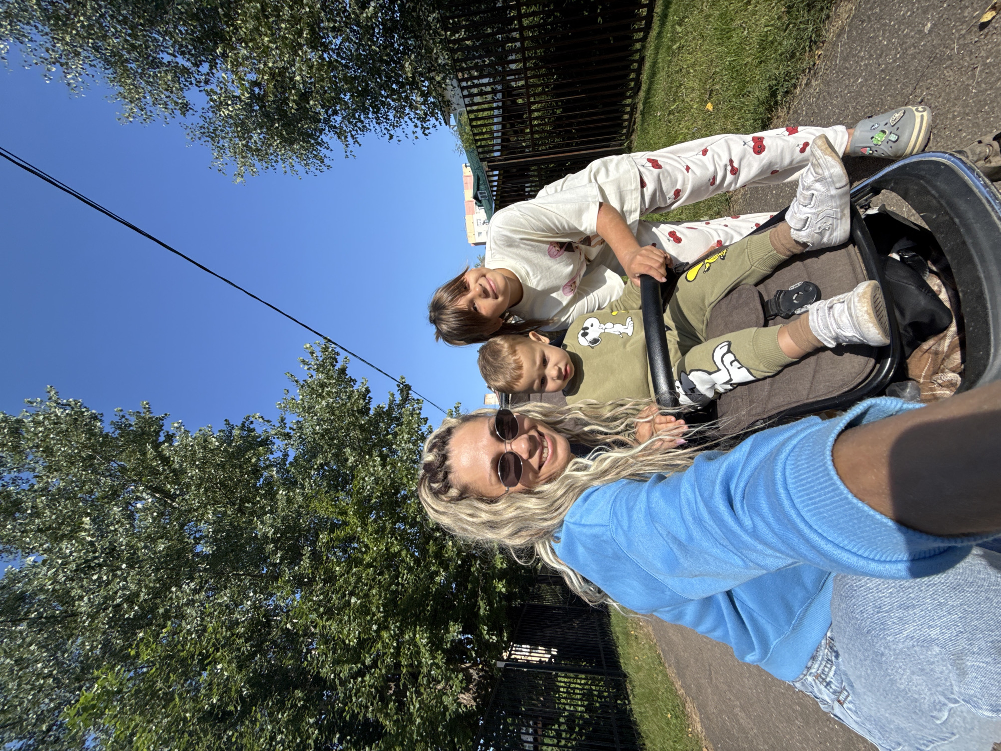 In Rusanovo, Russia, on a paved path bordered by grass and trees, a woman with blonde hair and sunglasses smiles at the camera while holding a stroller. A young boy sits in the stroller, wearing a green outfit with a Snoopy graphic. Beside him, a girl in a white t-shirt and patterned pants leans over the stroller, smiling. A black metal fence runs along the right side of the path, and a few buildings are visible in the background. The sky is clear and blue, and the sun is shining brightly.