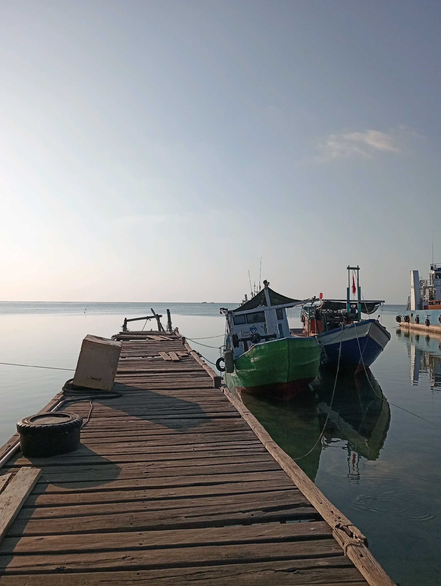 The image depicts a wooden pier extending into calm, reflective water. Two fishing boats are moored alongside the pier, one predominantly green and the other blue. A larger vessel, possibly a cargo ship, is visible in the background on the right. The sky is clear with a hint of haze. The pier itself shows signs of age and wear, with loose planks and weathered wood. On the pier, a black tire serves as a mooring or bumper, and a concrete block is positioned near its edge. The scene is set in Pagerungan Kecil, Indonesia, a location known for its fishing communities and coastal activities. The lighting suggests it is either early morning or late afternoon. There are no people visible in the image.