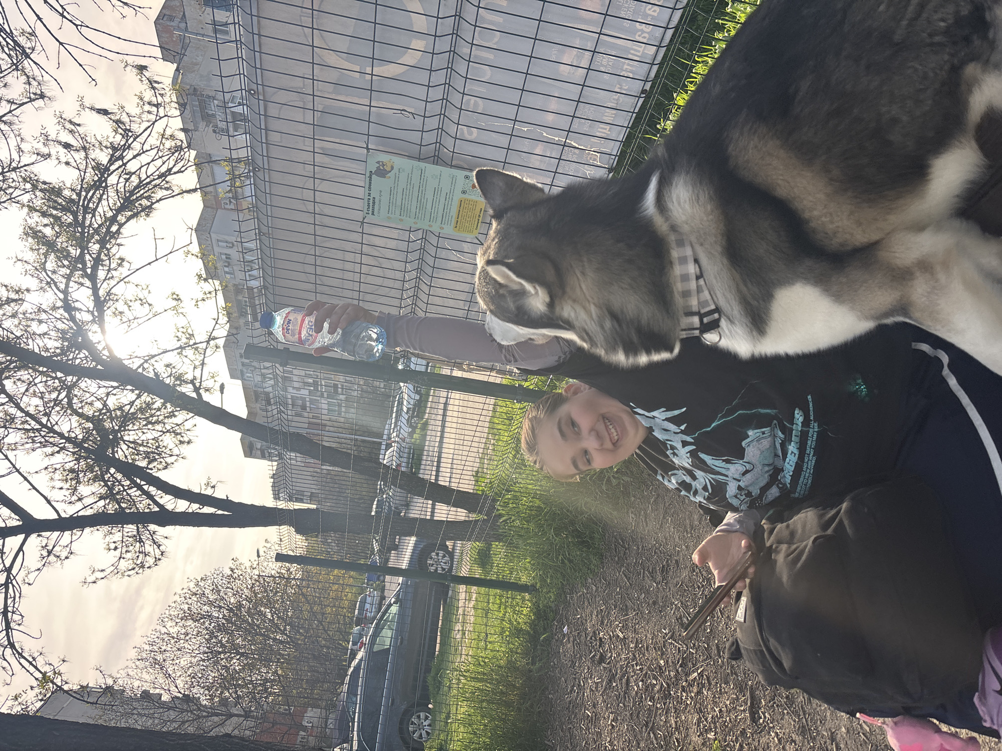 A person and a dog are captured in an outdoor setting. The person is smiling and appears to be interacting with the dog, which has a grey and white coat and is wearing a collar. A water bottle is visible in the background, held by a hand. The scene is partially obscured by a metal fence. Trees are present in the background.
The OCR indicates Bulgarian text, "5 съвета за спокойна разходка с плашливо куче," which translates to "5 tips for a calm walk with a fearful dog." The presence of Bulgarian text and a "Devin" water bottle suggests the location is in Bulgaria. Considering the buildings and vegetation visible, a park or green space in a city is likely. Without additional information, definitively identifying the exact location in Plovdiv is not possible, but it's plausible the scene is in a park or dog-friendly area within the city.