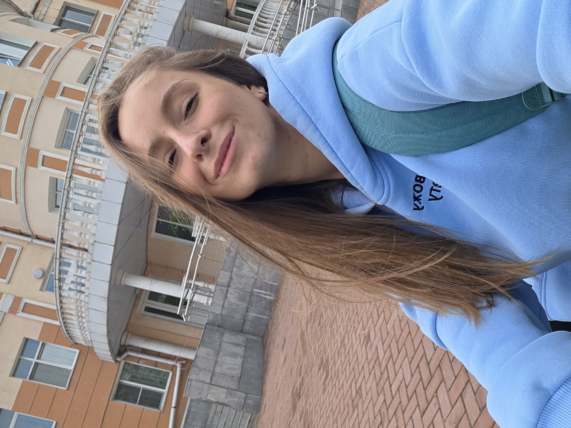 A young woman is taking a selfie outdoors. She's wearing a light blue hoodie and a green backpack, and is standing in front of a brick building with light-colored columns and detailed trim. The ground is paved with red bricks. The text on the hoodie, "УЛА ВОХА," is partially visible. The location is Birobidzhan, Russia.