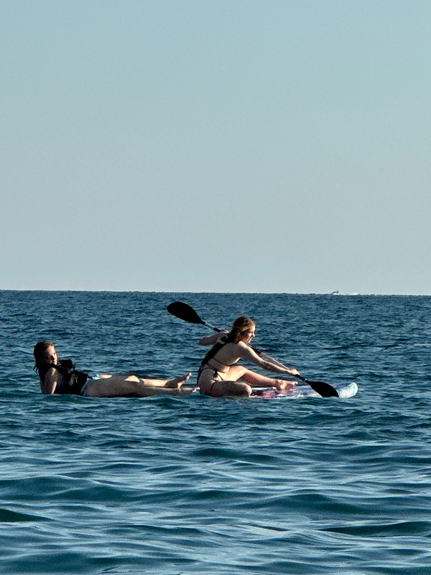Two young women are paddleboarding on a calm, blue ocean under a clear, pale blue sky. The woman in the foreground is seated and actively paddling, wearing a bikini. The woman in the background is lying on her stomach on the paddleboard, also wearing a bikini. Both are on the same paddleboard. The water is choppy and reflects the sky. In the distance, a faint outline of a boat can be seen on the horizon. The scene appears to be a recreational activity in a serene marine environment. The location is the sea off the coast of Boreti, Montenegro.