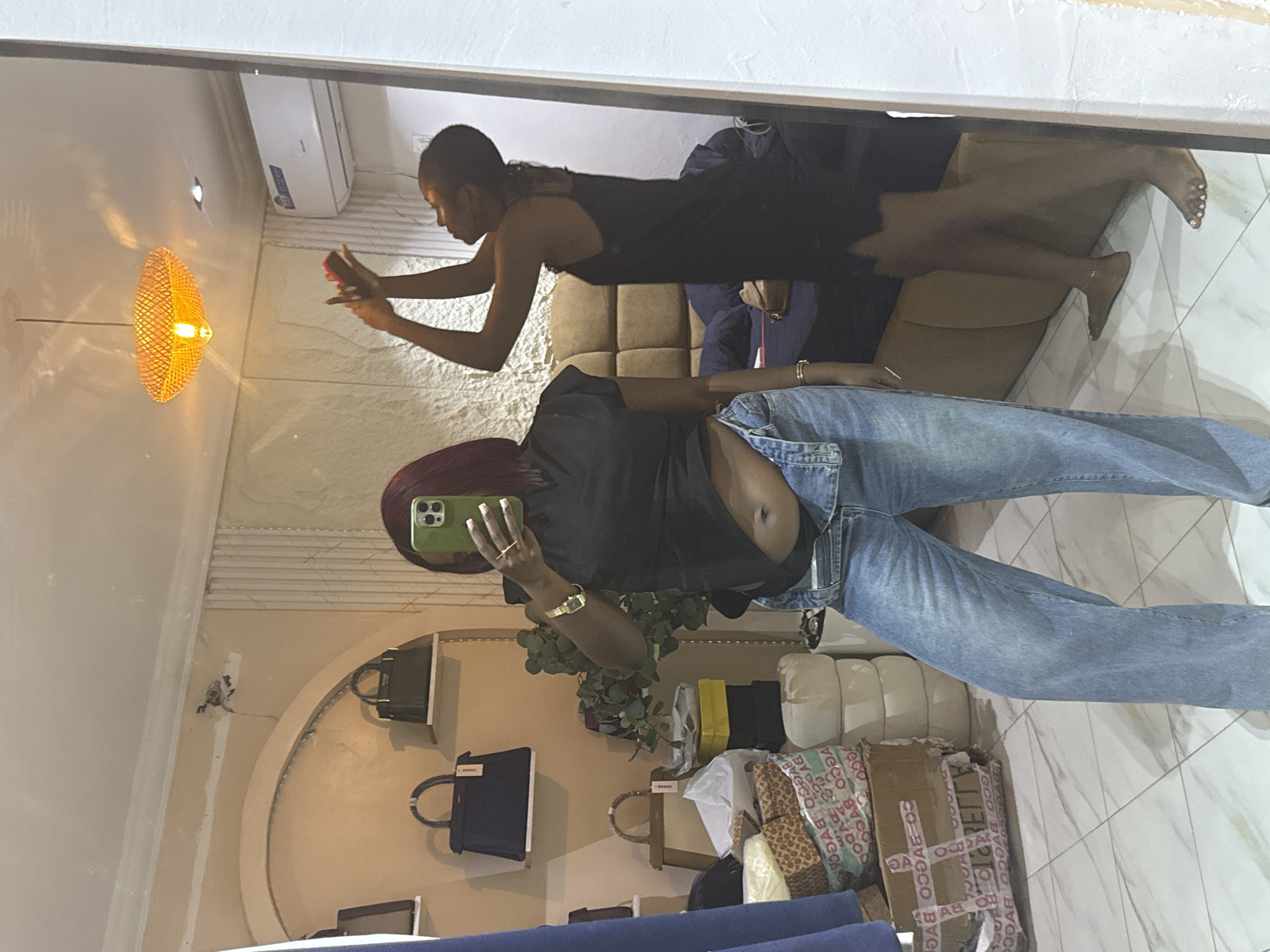 In a room, likely a bedroom or dressing room, two women are visible in a mirror's reflection. The room has tiled flooring and pale walls. To the left, a woman with burgundy hair is taking a selfie in a mirror. She is wearing a black t-shirt with rolled-up sleeves and light blue wide-leg jeans, with the top of her jeans unbuttoned. She is also wearing a gold watch and a ring on her left hand. Behind her, shelves display several handbags. In the foreground, stacks of boxes and some decorative items are visible.
To the right of the first woman, another woman is reflected in the mirror. She is wearing a black slip dress and is reaching up towards the ceiling, seemingly adjusting a light fixture or interacting with an object above. She is barefoot. A bed with a brown quilted headboard is behind her. A white air conditioning unit is mounted on the wall. The overall impression is an indoor setting, possibly a residential space, in Abuja, Nigeria.