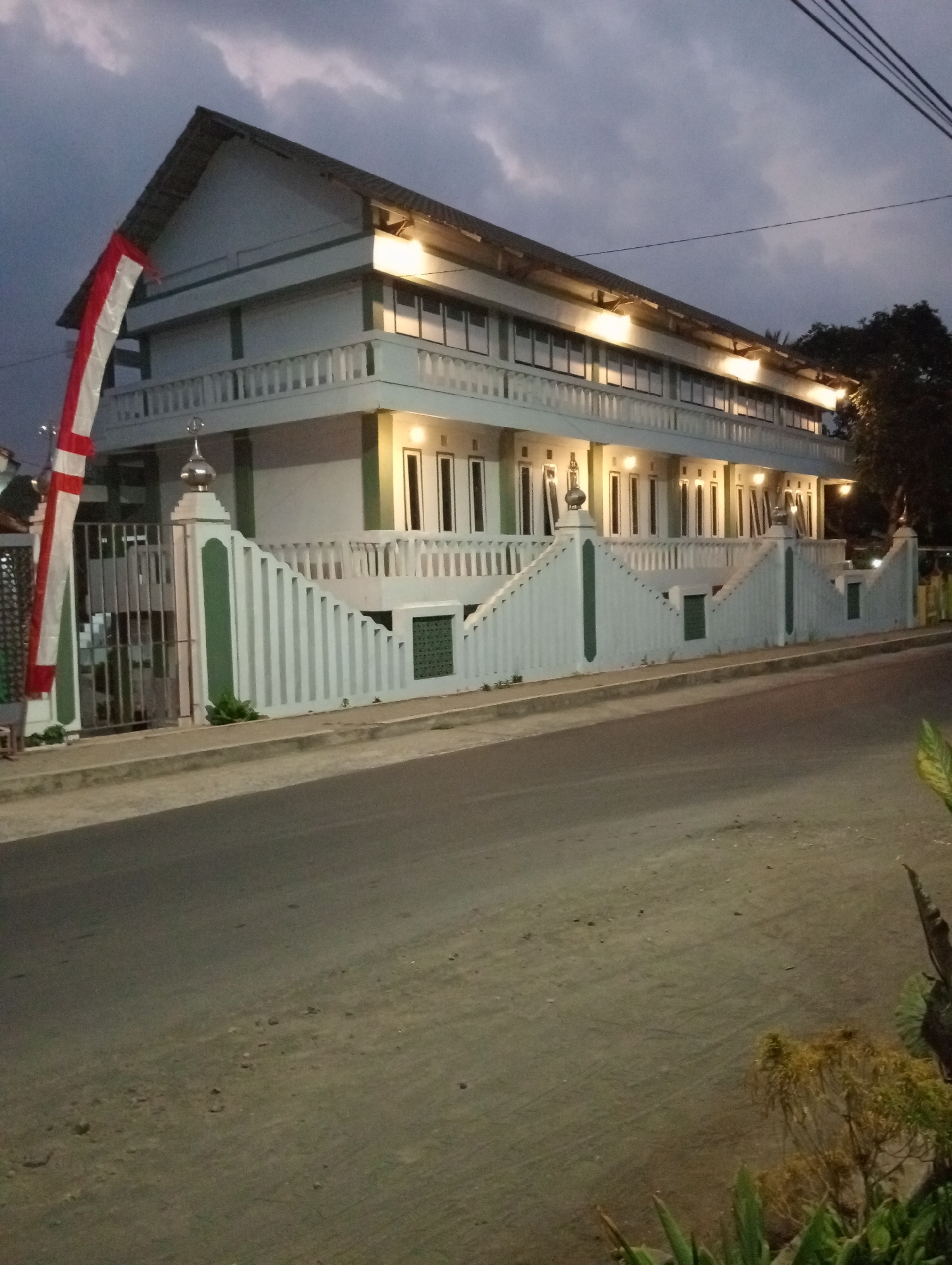 The image depicts a multi-story building with a white facade and green trim, located in Desa Silo, Indonesia. The building appears to be a religious or community center, possibly a mosque, given its architectural style and the presence of decorative elements like the finial on the gatepost. The building has multiple windows and a balcony on the upper floor, and is illuminated by artificial lights, suggesting it is evening or nighttime. In front of the building is a white, decorative fence with green accents, and a metal gate. A red and white flag, likely the Indonesian flag, is hung vertically on the left side of the building. The building is situated next to a paved road, with some vegetation visible in the foreground and to the right. The sky above is cloudy and dark, indicating dusk or a stormy weather condition.